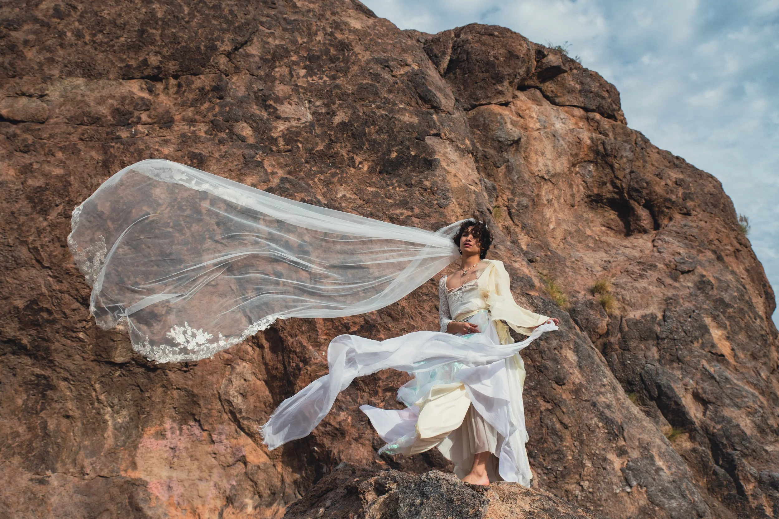 A woman in a flowing, white and cream-colored gown stands among large rocks with a vast sky overhead, her veil flowing in the wind.