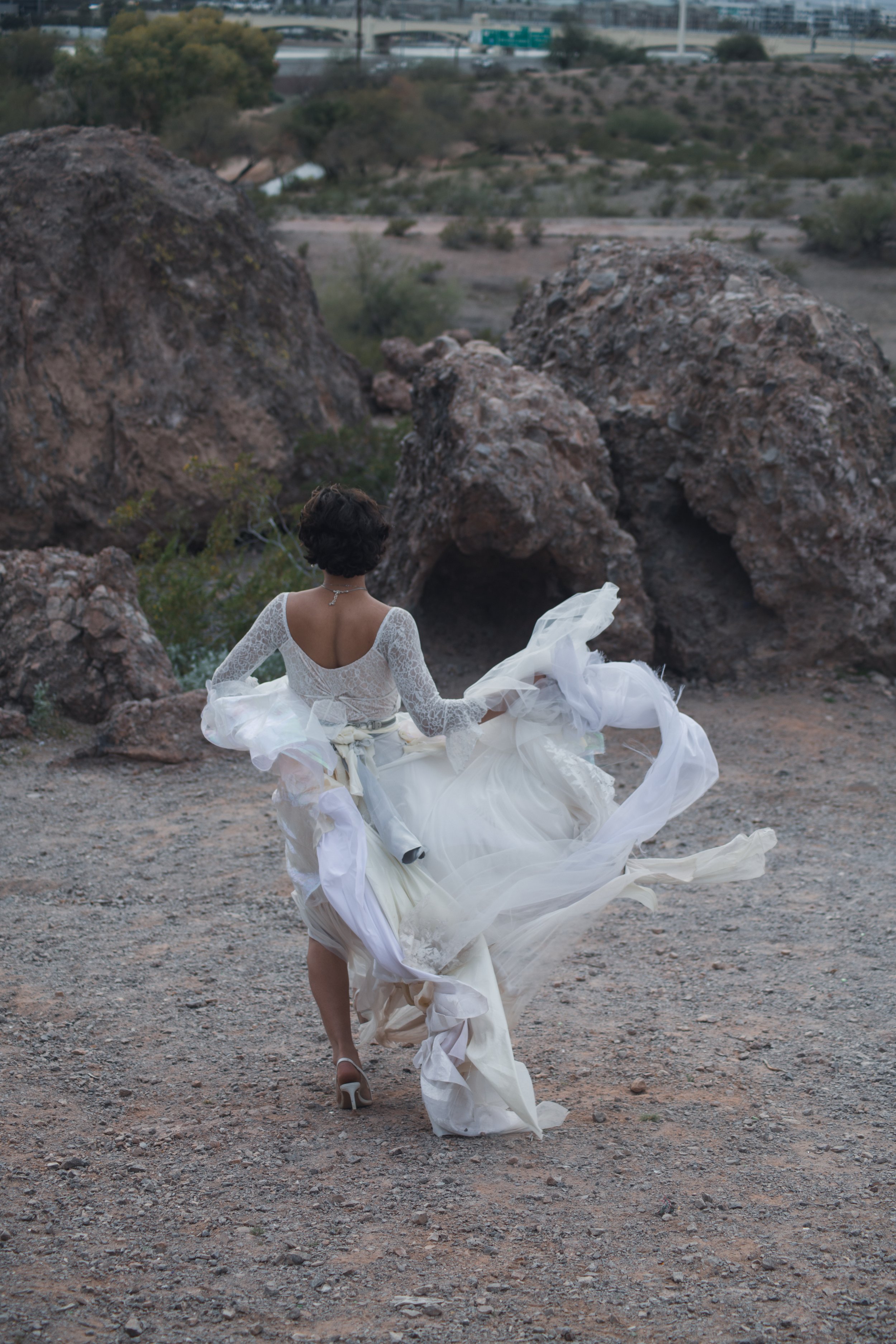 A woman in a white wedding dress running outdoors in a desert-like landscape with large rocks, with her back facing the camera.