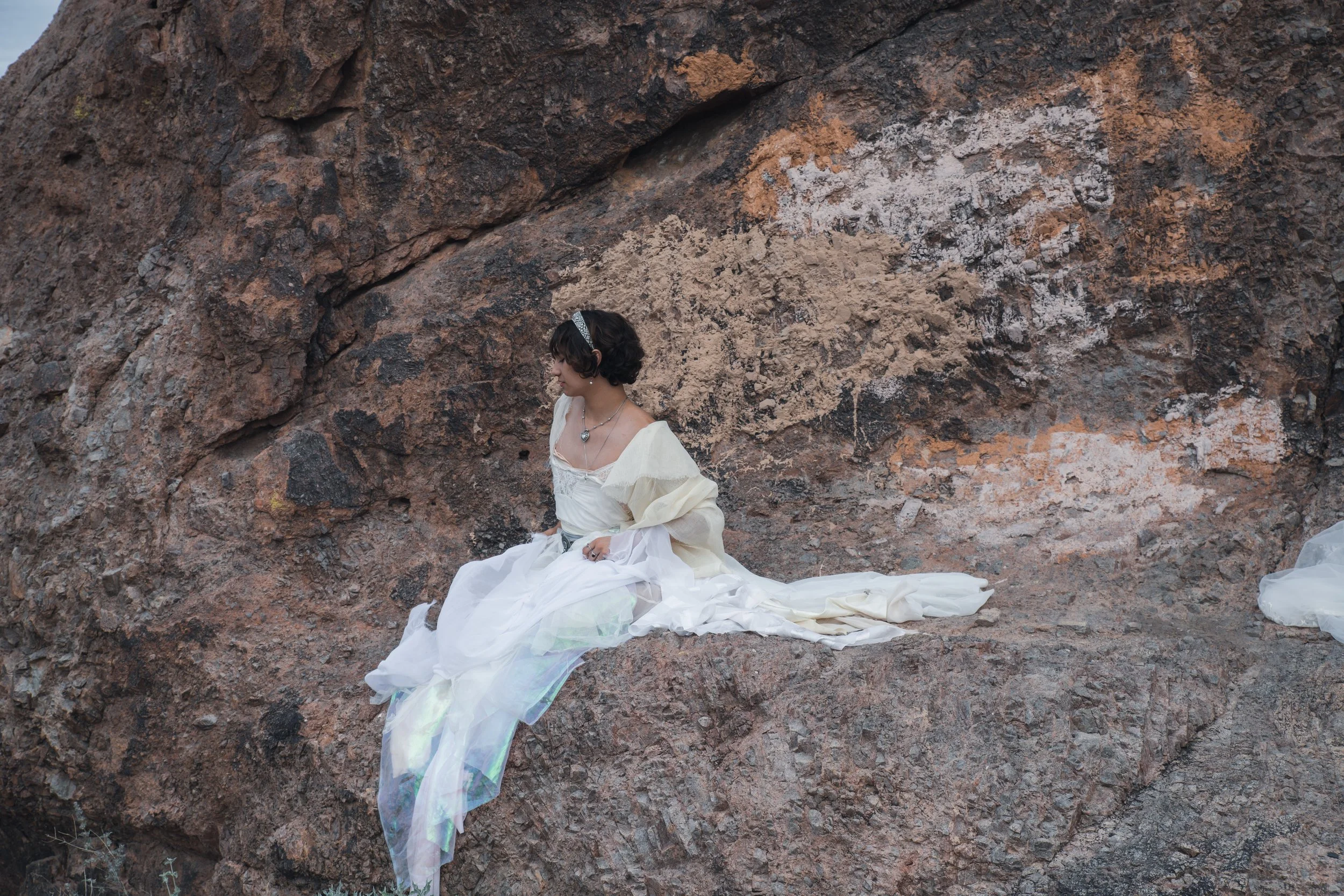 A woman in a white dress with a heart-shaped necklace sits against a large rocky wall.