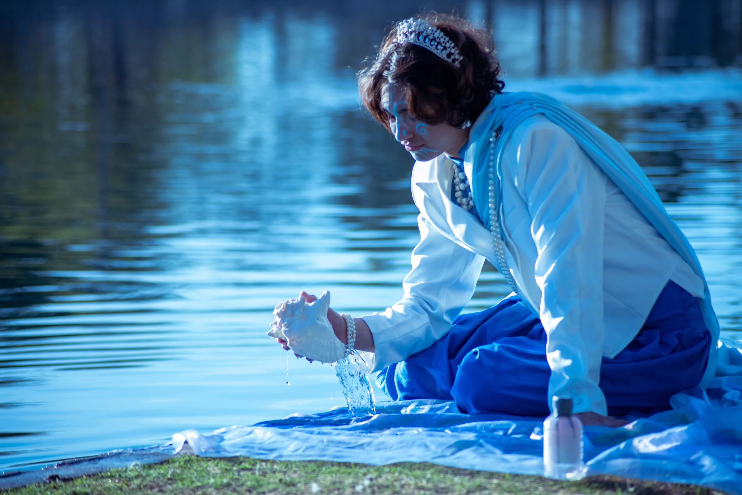 Woman in white dress and tiara kneeling by a lake, washing or rinsing fabric or clothing.