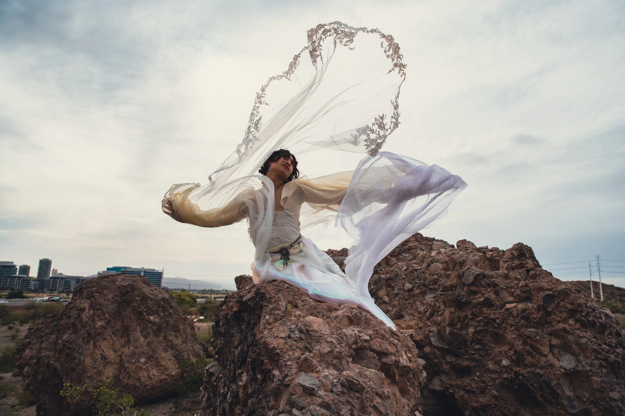 A woman in a flowing dress standing on a rocky hilltop with a cityscape and cloudy sky in the background.