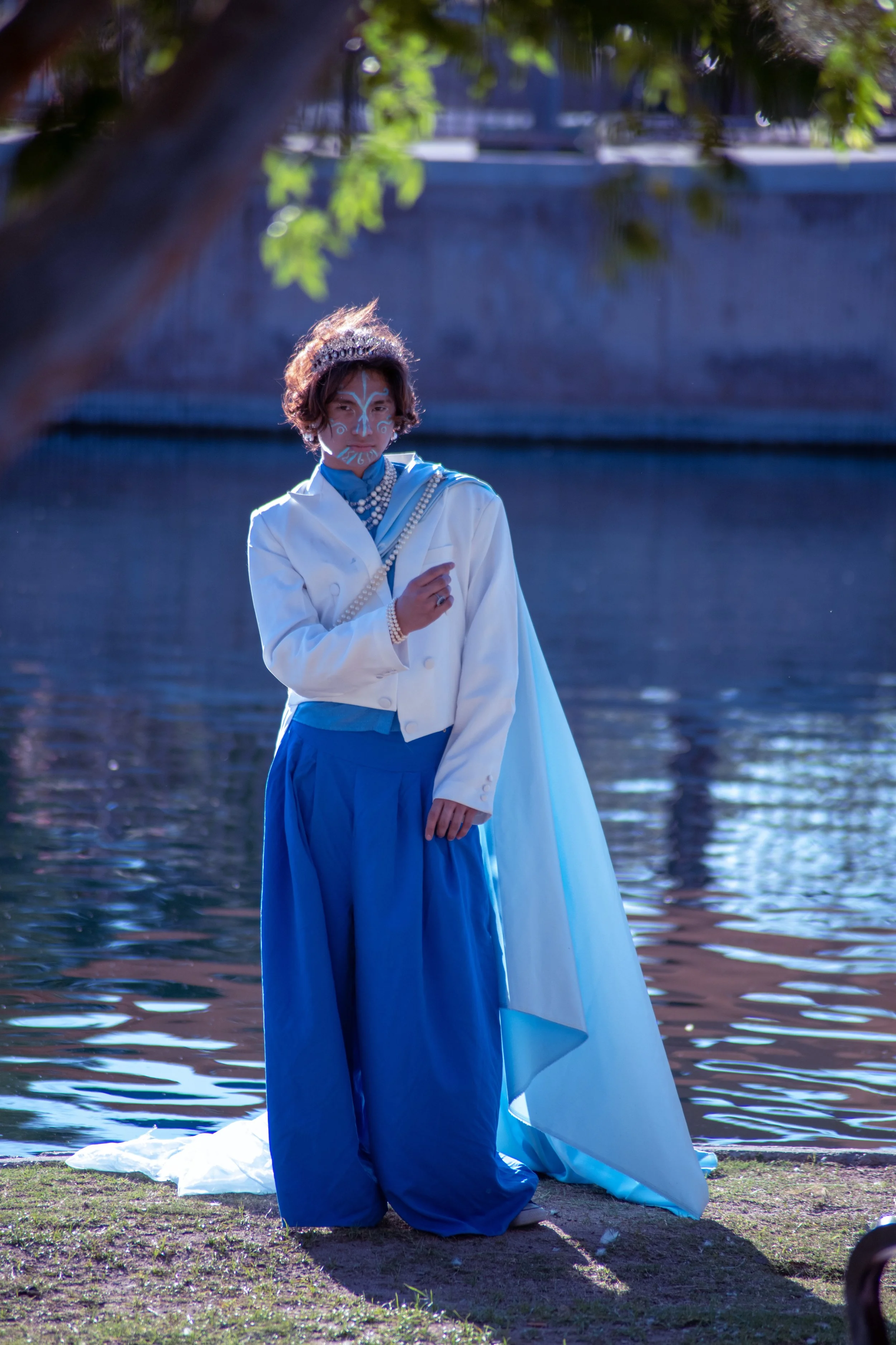Person with face paint and a crown standing by the water, dressed in blue and white cloak, with a large white and blue fabric train behind them, outdoor setting.