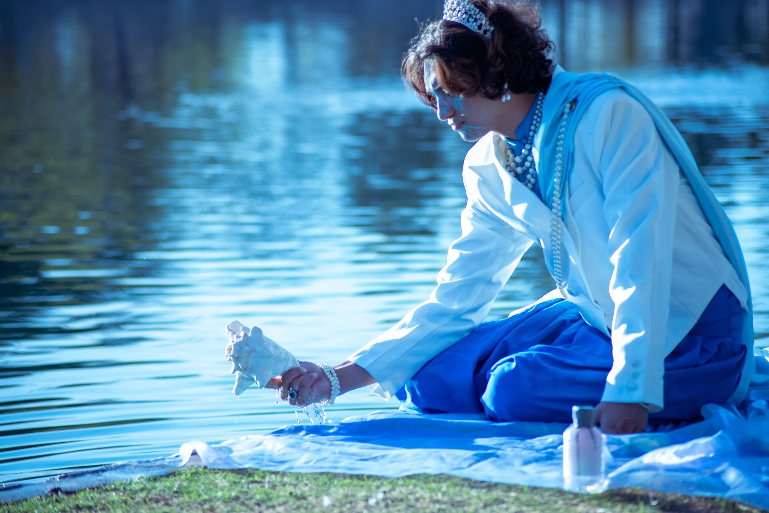 A man dressed in white with a tiara and pearl jewelry is kneeling by a body of water, holding and examining a seashell.