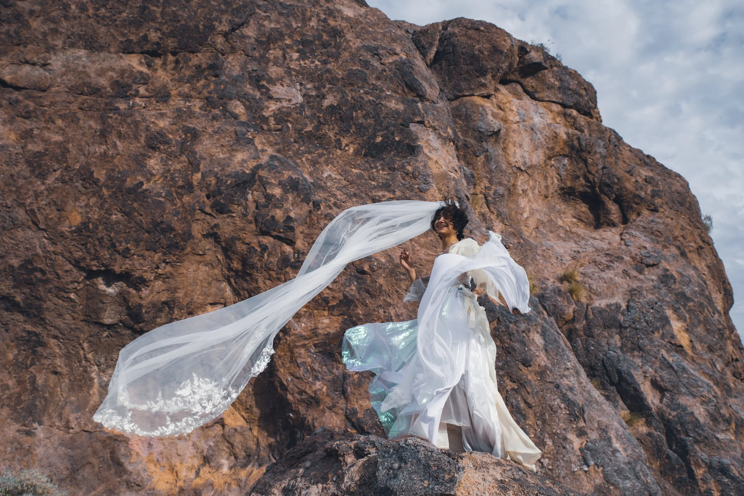 A woman in a white dress with long flowing fabric stands on rocks by a large rocky cliff, smiling with her hair blowing in the wind, against a partly cloudy sky.