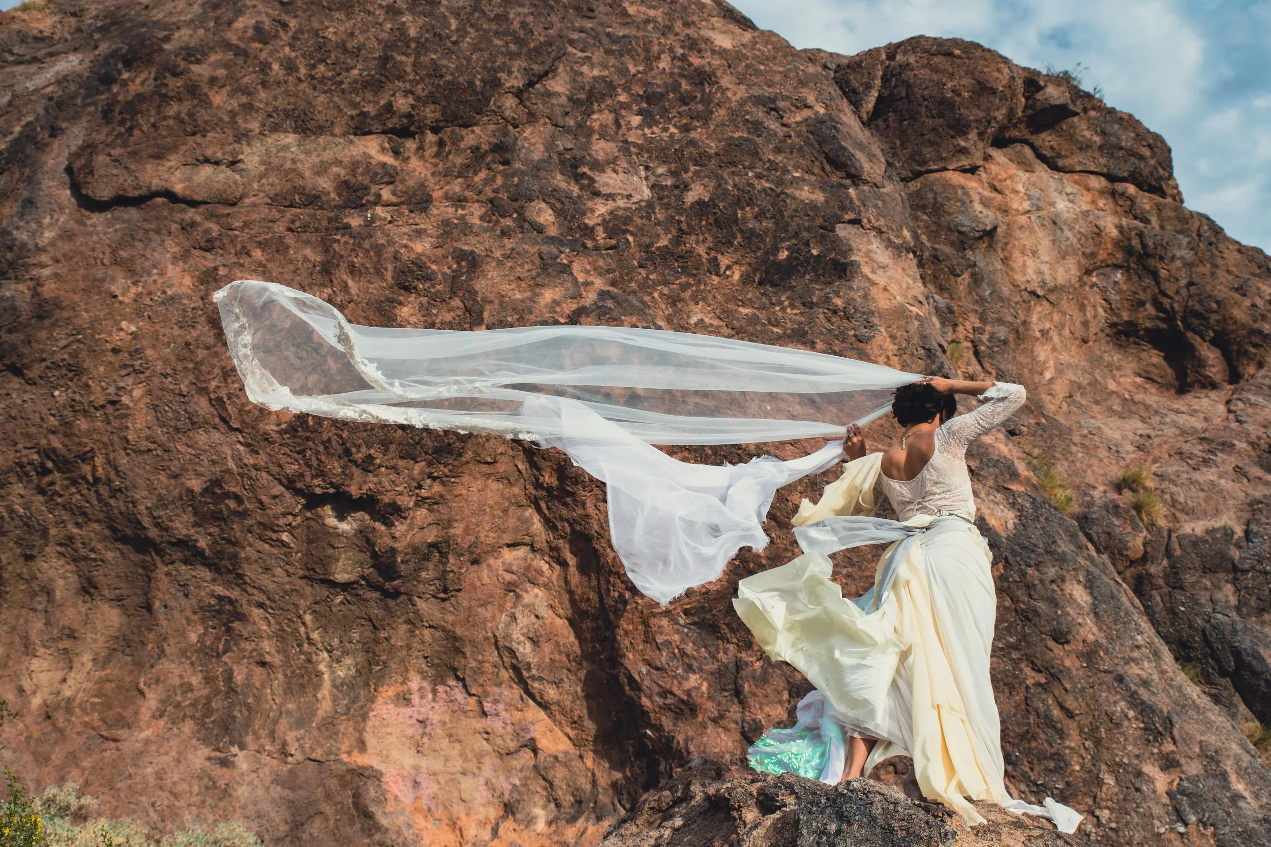 A person dressed in a white costume with long flowing fabric, sitting on rocky terrain against a backdrop of reddish-brown cliffs under a partly cloudy sky.