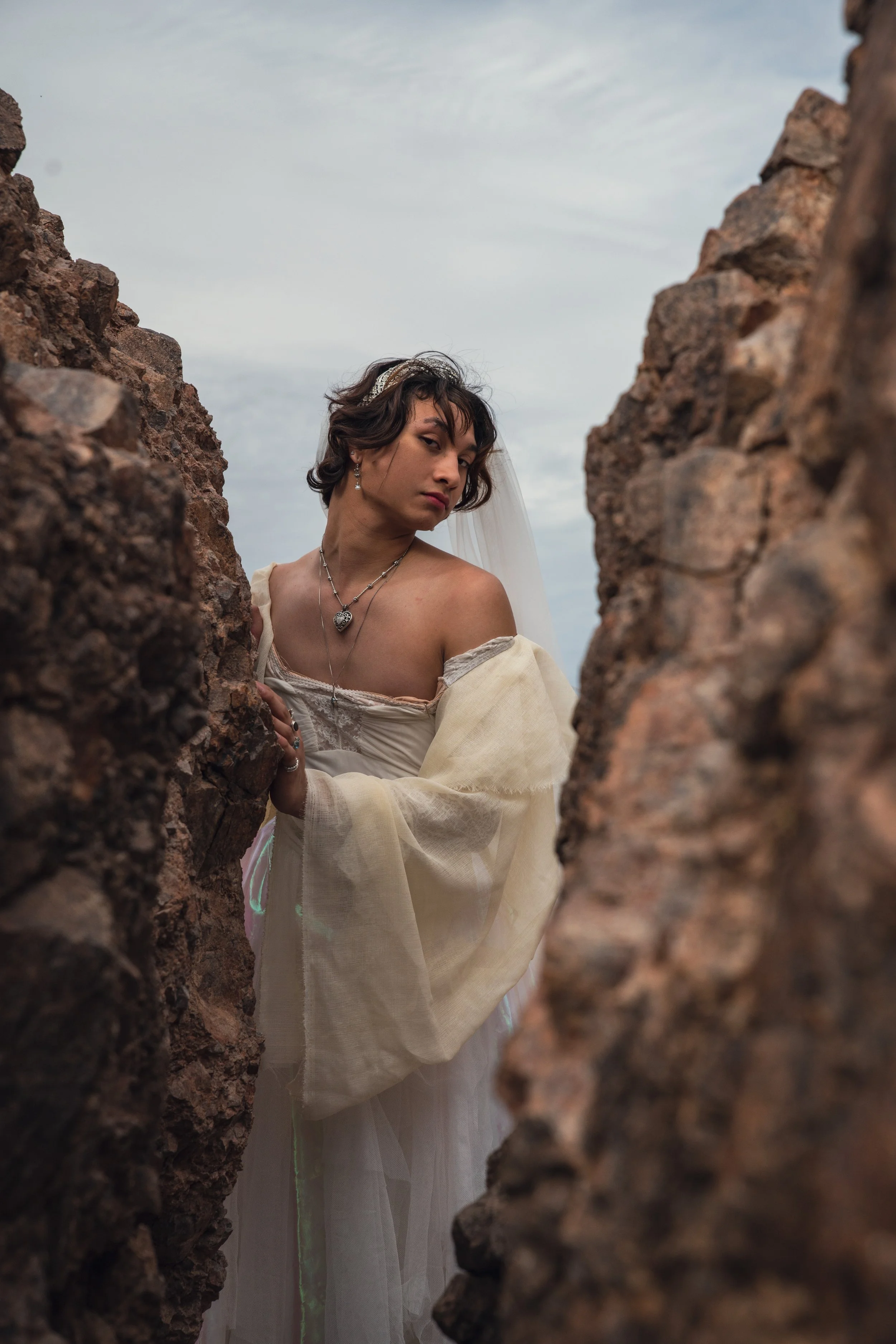 A man with short dark curly hair wearing a vintage-inspired off-white dress with a sheer puffed sleeve, adorned with layered silver jewelry, including a necklace and earrings. She is standing outdoors between two large, rugged rocks, with a cloudy sk
