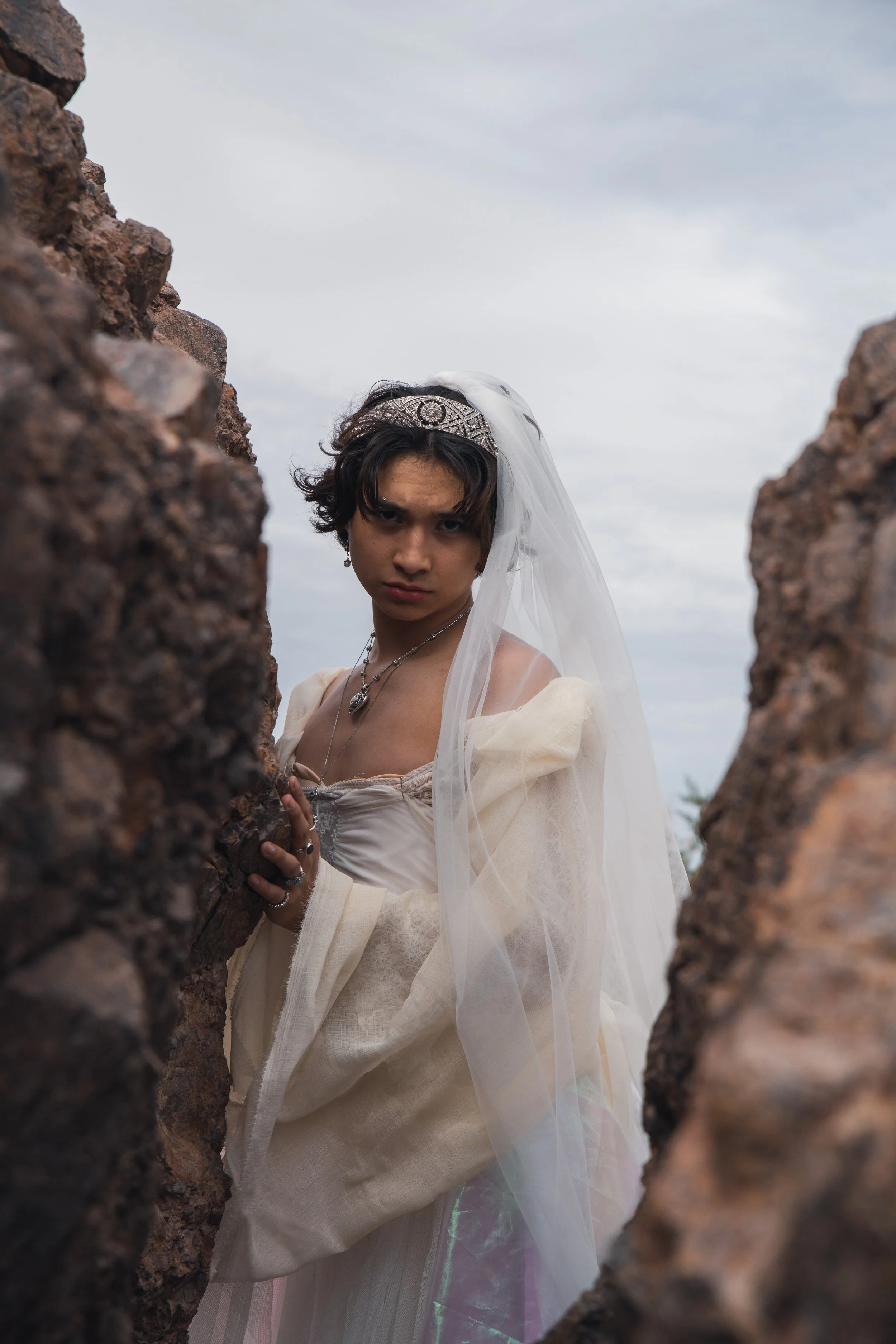 Young person dressed in wedding attire, wearing a tiara and a veil, standing between rocks outdoors under cloudy sky.