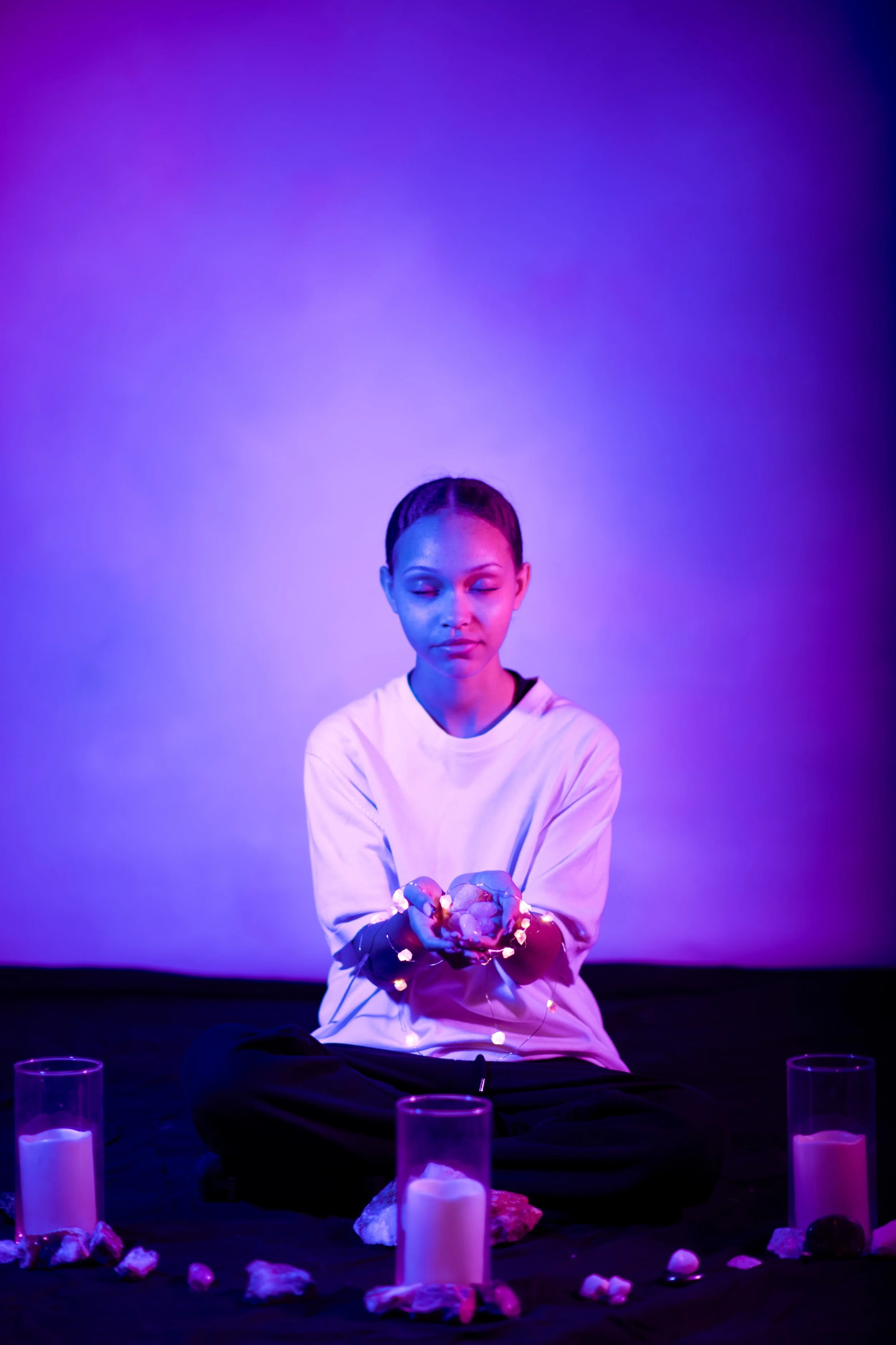 A person with braided hair is sitting cross-legged in a dark room illuminated by purple and blue lights. They are wearing a white t-shirt, holding a bunch of stones and fairy lights in their hands. Around them are candles and rocks on a black surface.