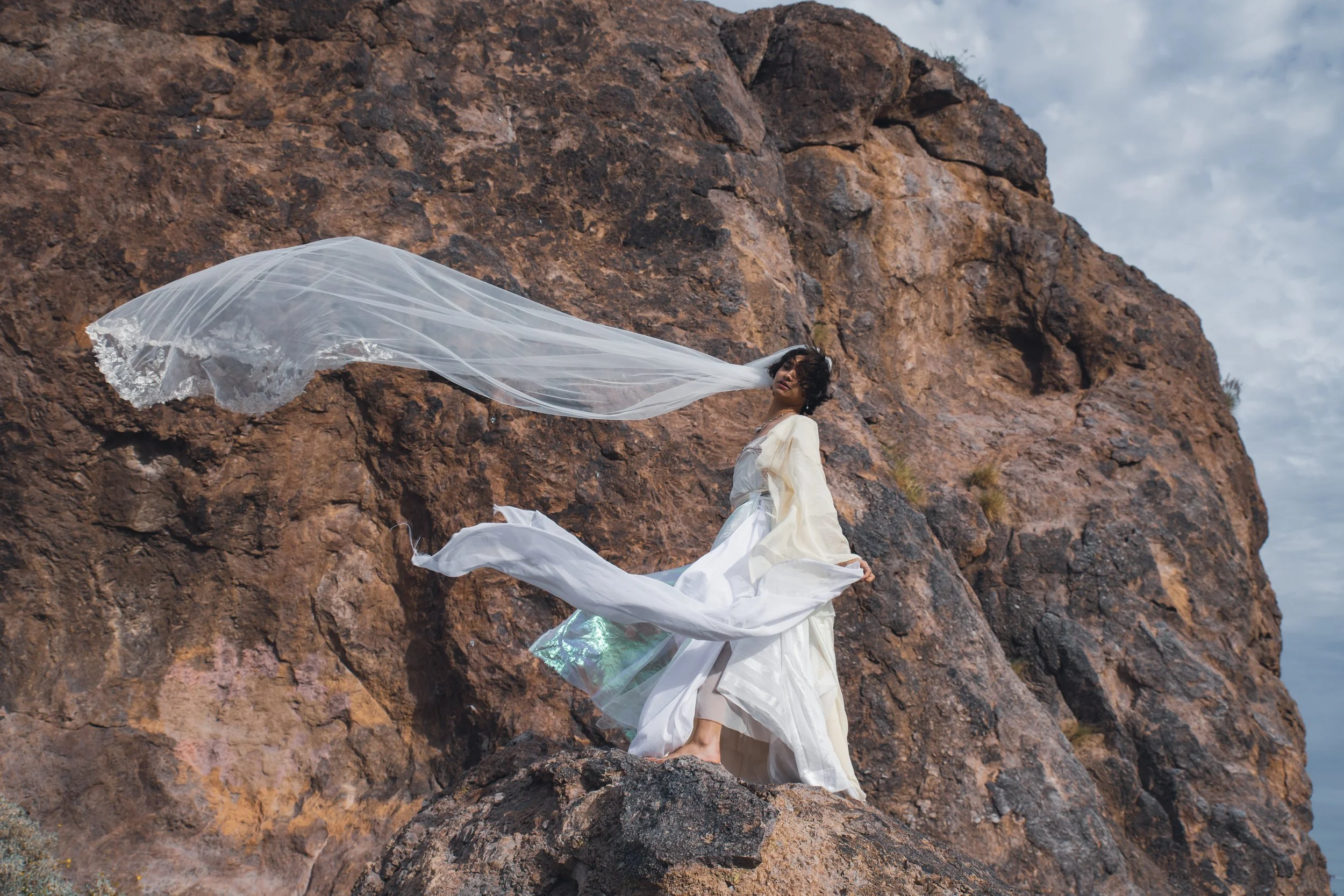 A woman dressed in a flowing white gown stands among large rocks, with a sheer white fabric billowing in the wind against a backdrop of a cloudy sky.