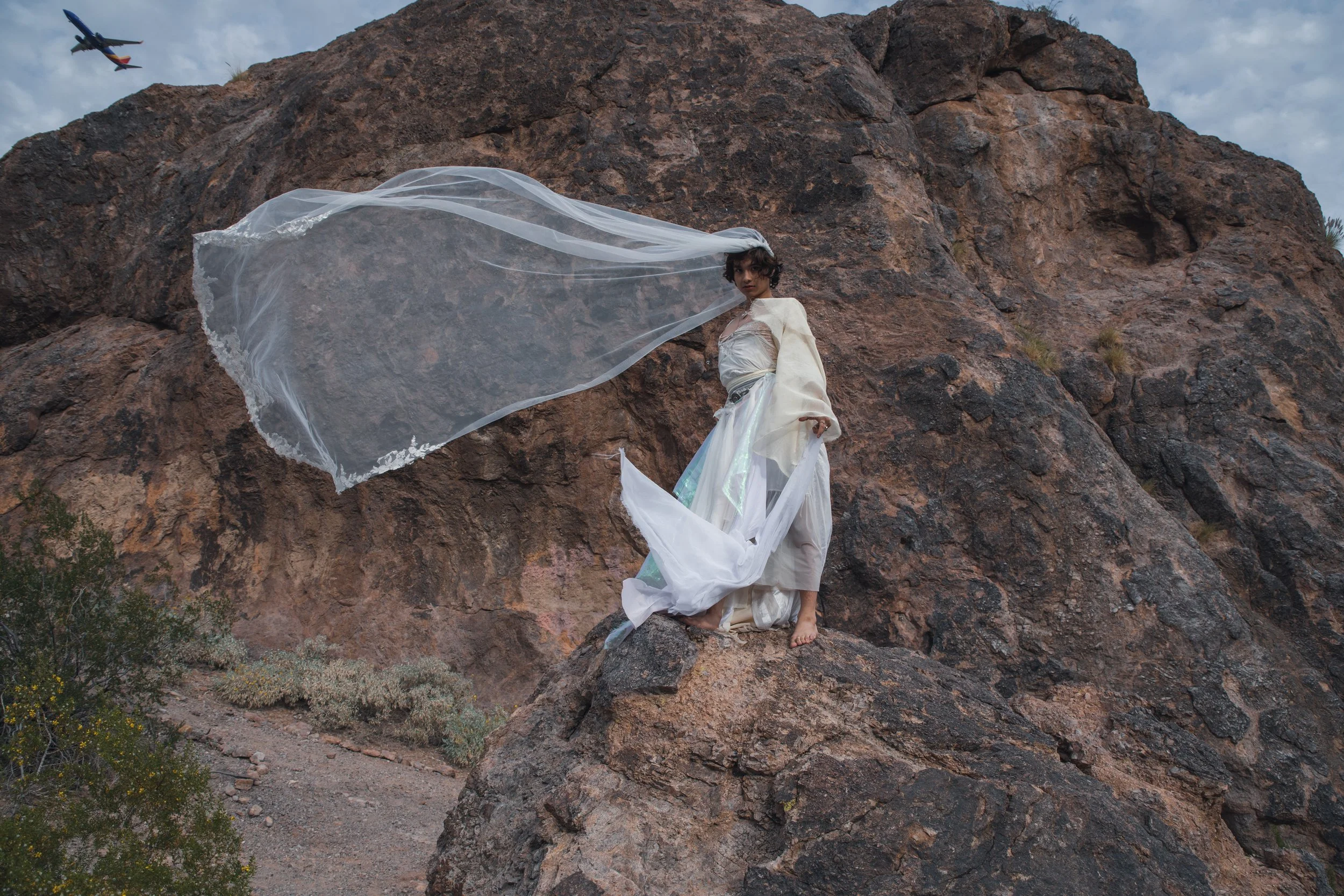 A woman dressed in white flowing garments standing on a rocky hillside with a mountain behind her, a large transparent fabric flowing in the wind, and an airplane flying in the cloudy sky.