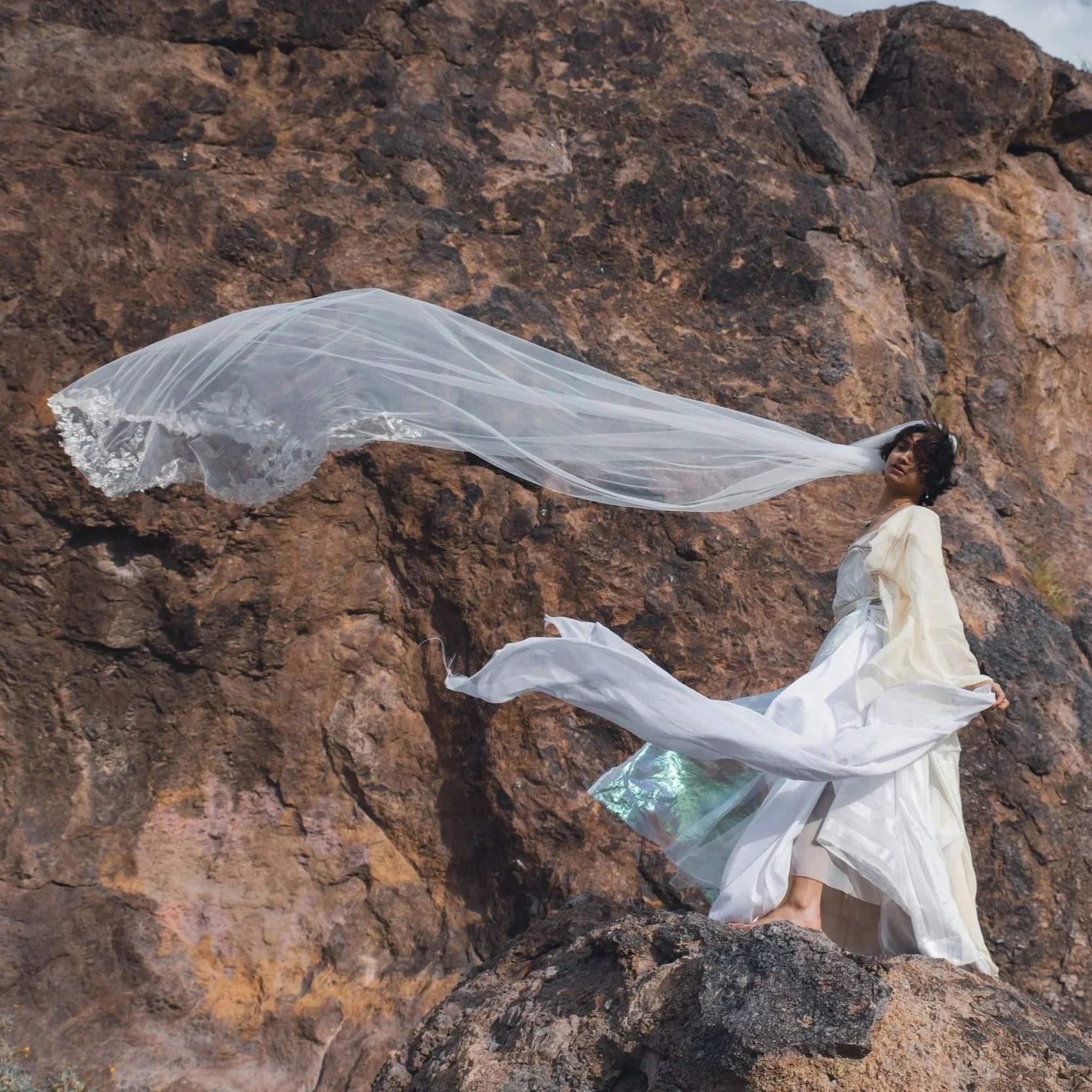 A person dressed in flowing white clothing and a veil is standing on rocky terrain near a large rock formation. Their hair is short and dark, and they are facing towards the camera with a neutral expression. The veil is billowing in the wind.