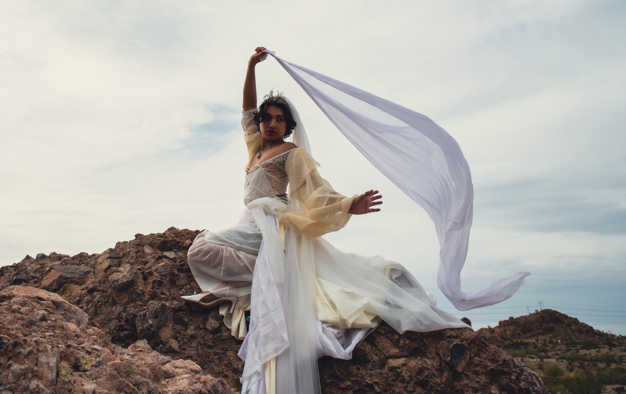 A person dressed in a flowing white and beige costume, sitting on rocky terrain outdoors during daytime, holding a long white fabric that billows in the wind against a partly cloudy sky.