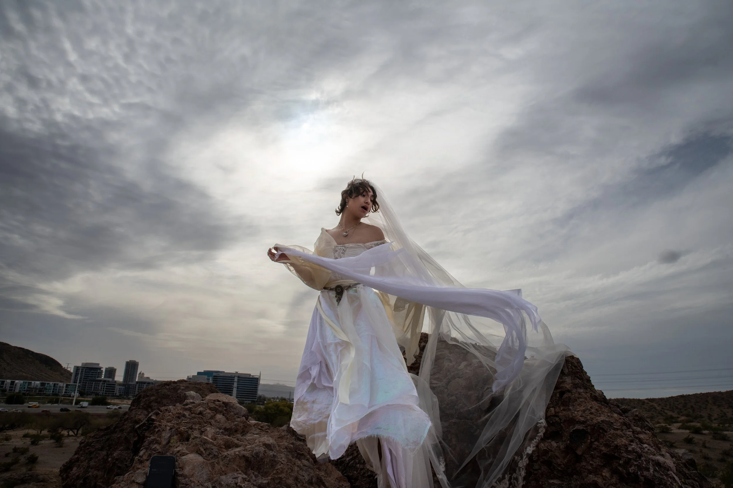 A man in a white dress standing on rocks outdoors under a cloudy sky with city buildings in the background.