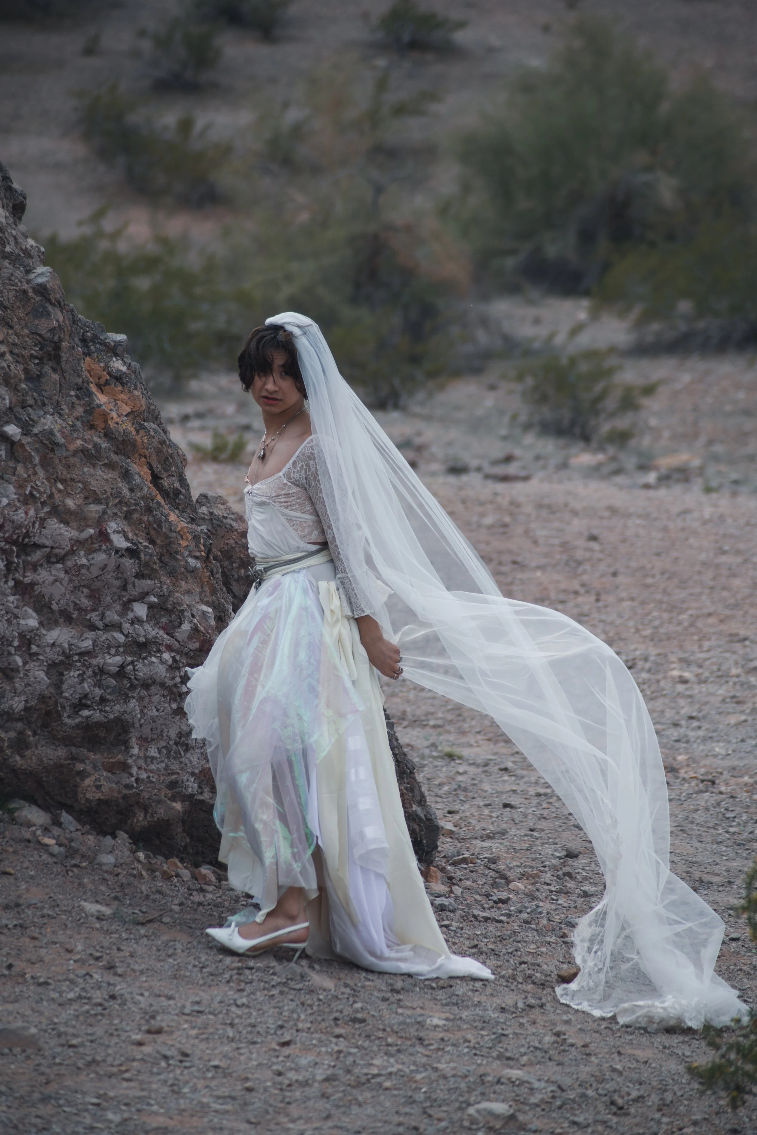 A woman dressed in a wedding gown, standing outdoors on rocky terrain, with a veil and white high heels, looking at the camera.