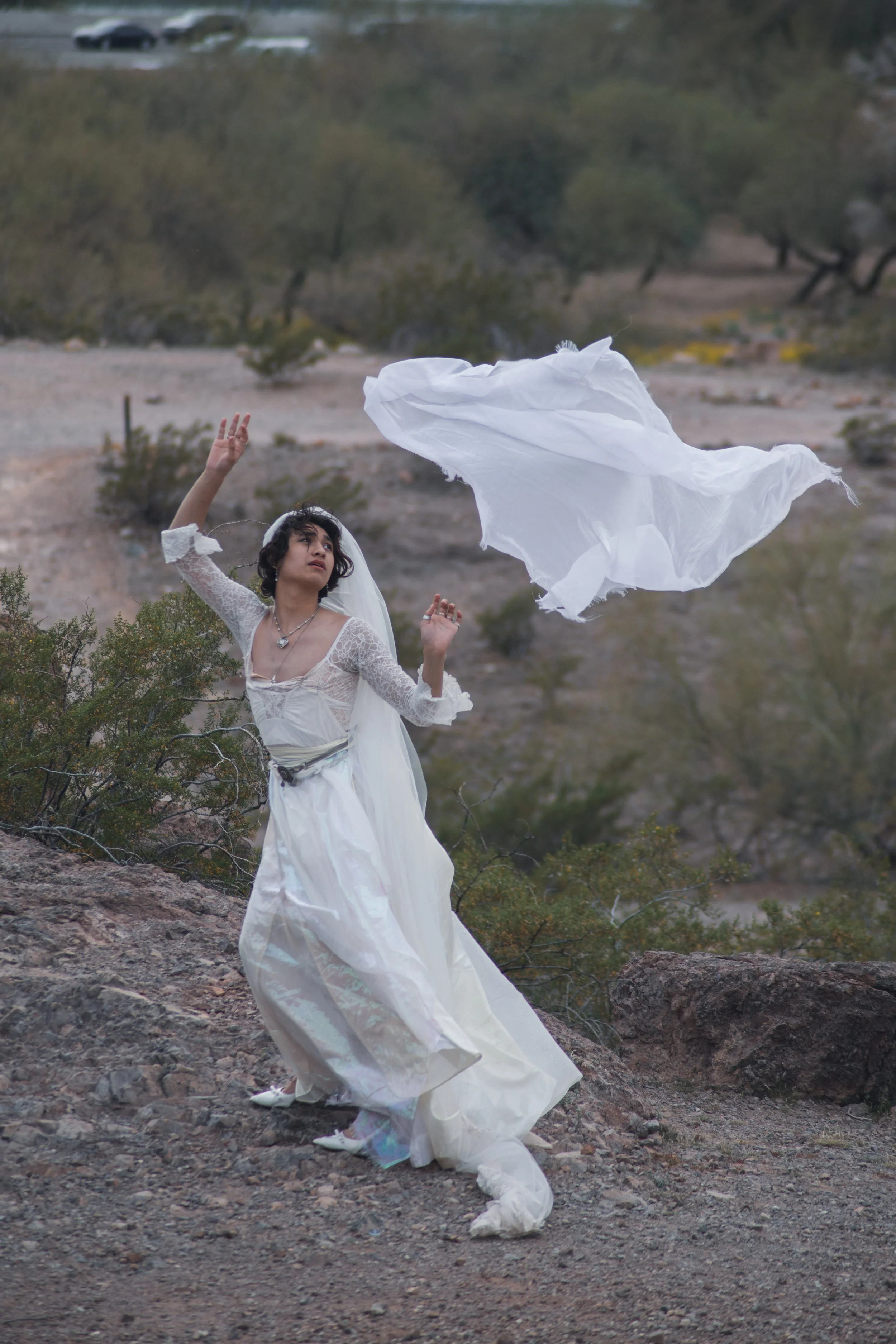A woman in a white dress and veil standing outdoors on rocky terrain, with a large white fabric flowing in the wind, under an overcast sky.