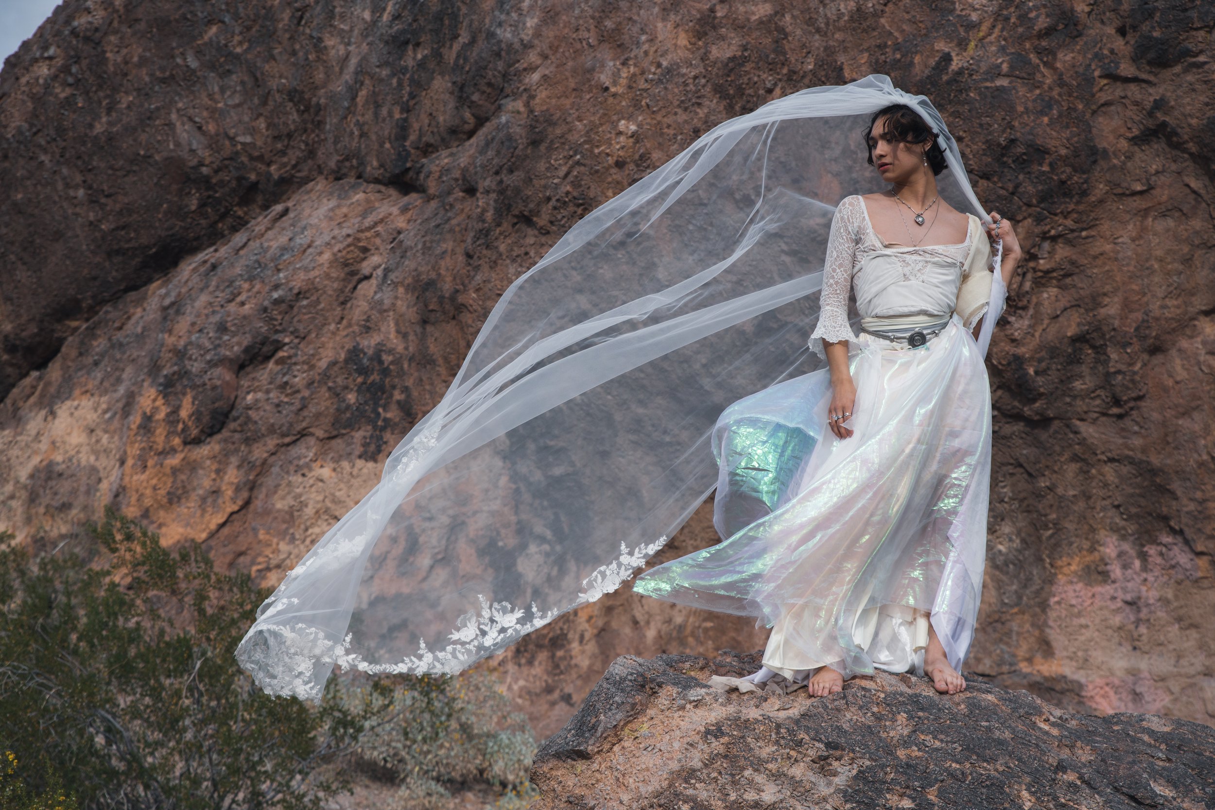Woman in a white dress with a flowing veil standing on a rock in front of a rocky mountain.