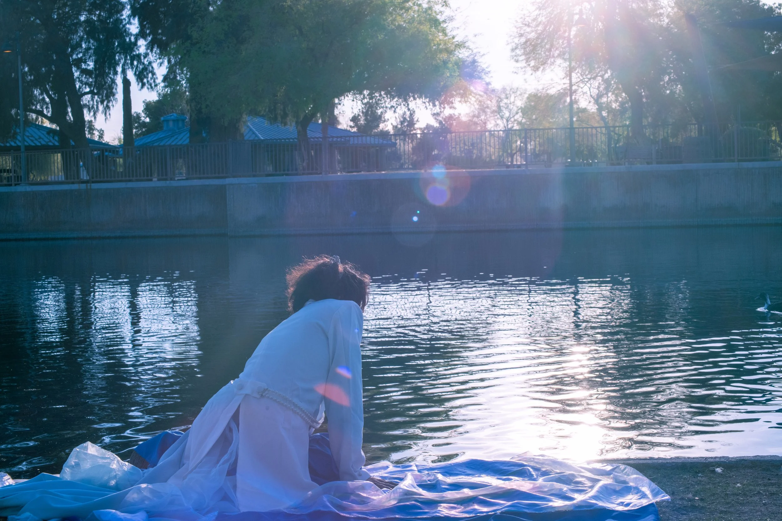 Person in white clothing sitting by a body of water with trees and sunlight in the background.
