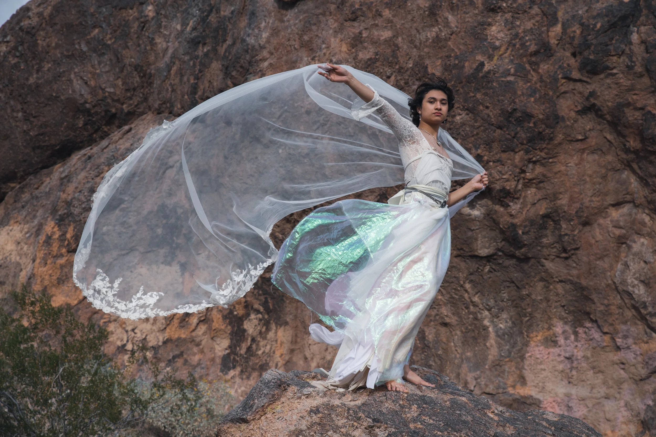 A young man with short dark hair and light skin stands on a rock in front of a rocky mountain background. He wears a white lace long-sleeve top and a flowing iridescent skirt. He holds a sheer, flowing veil that billows in the wind.