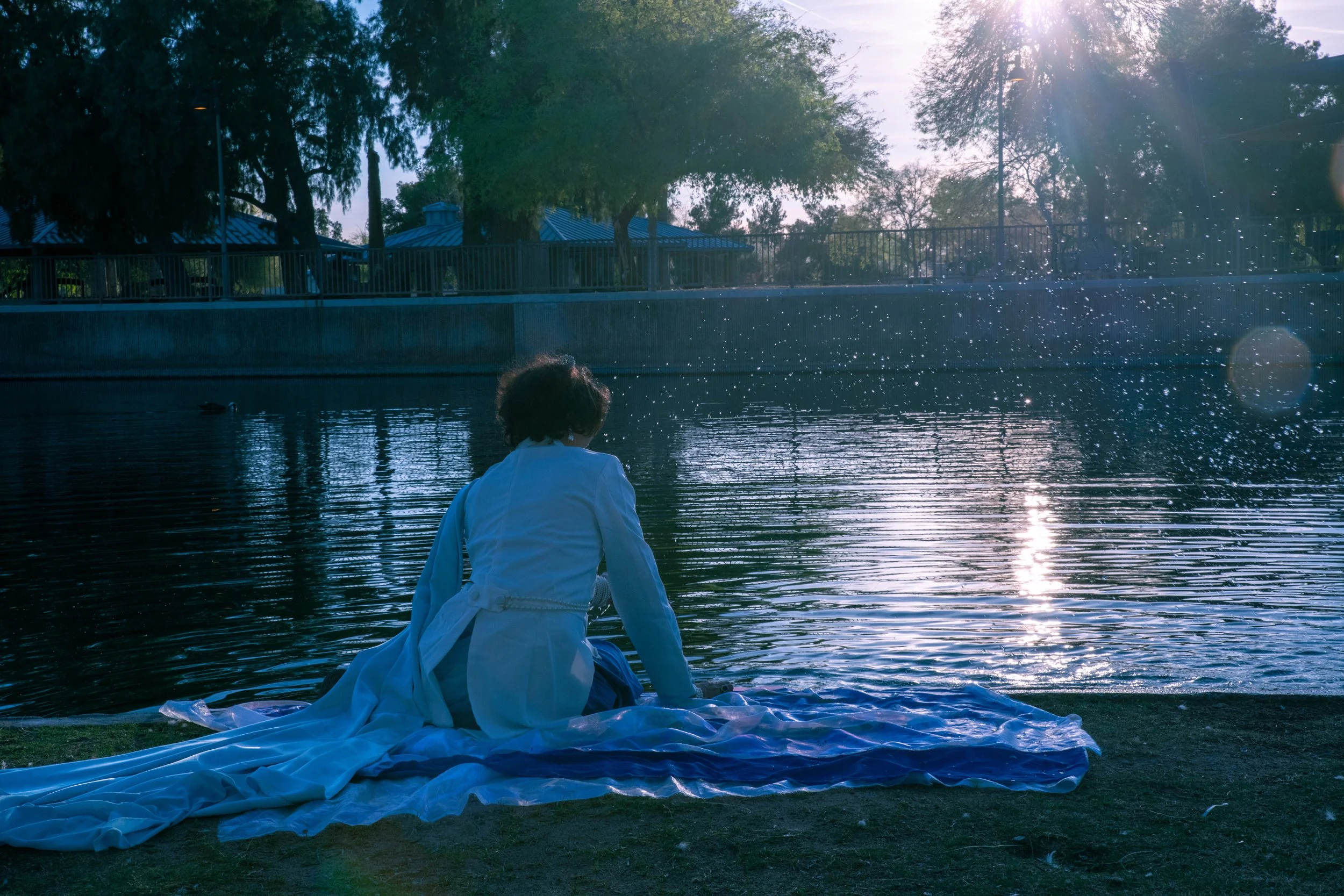 A person dressed in white, sitting by a body of water with a white cloth extended on the ground, overlooking the water at sunset or sunrise with trees and a blue-roofed structure in the background.