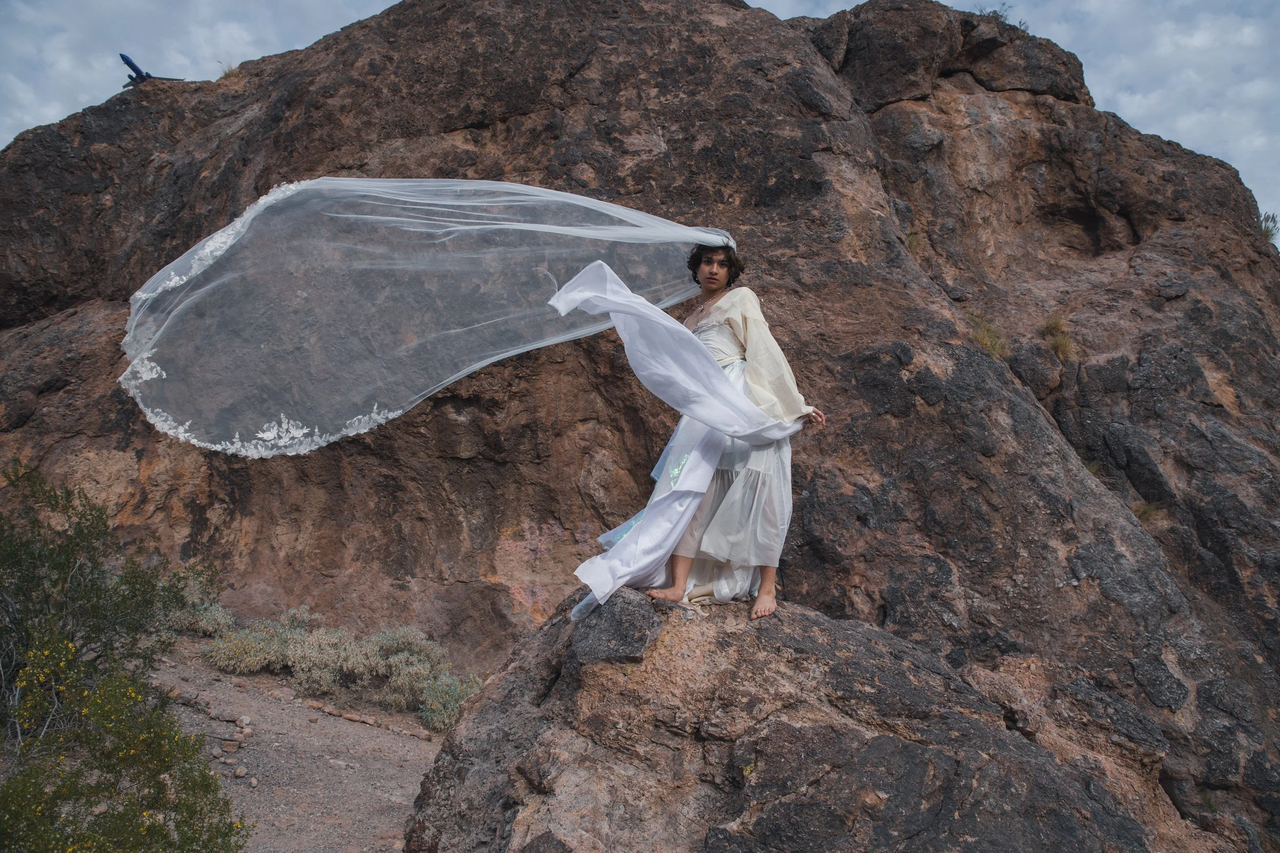 A person dressed in white is standing on a large rock formation outdoors, with a billowing sheer fabric flowing in the wind and a small airplane visible in the sky above.