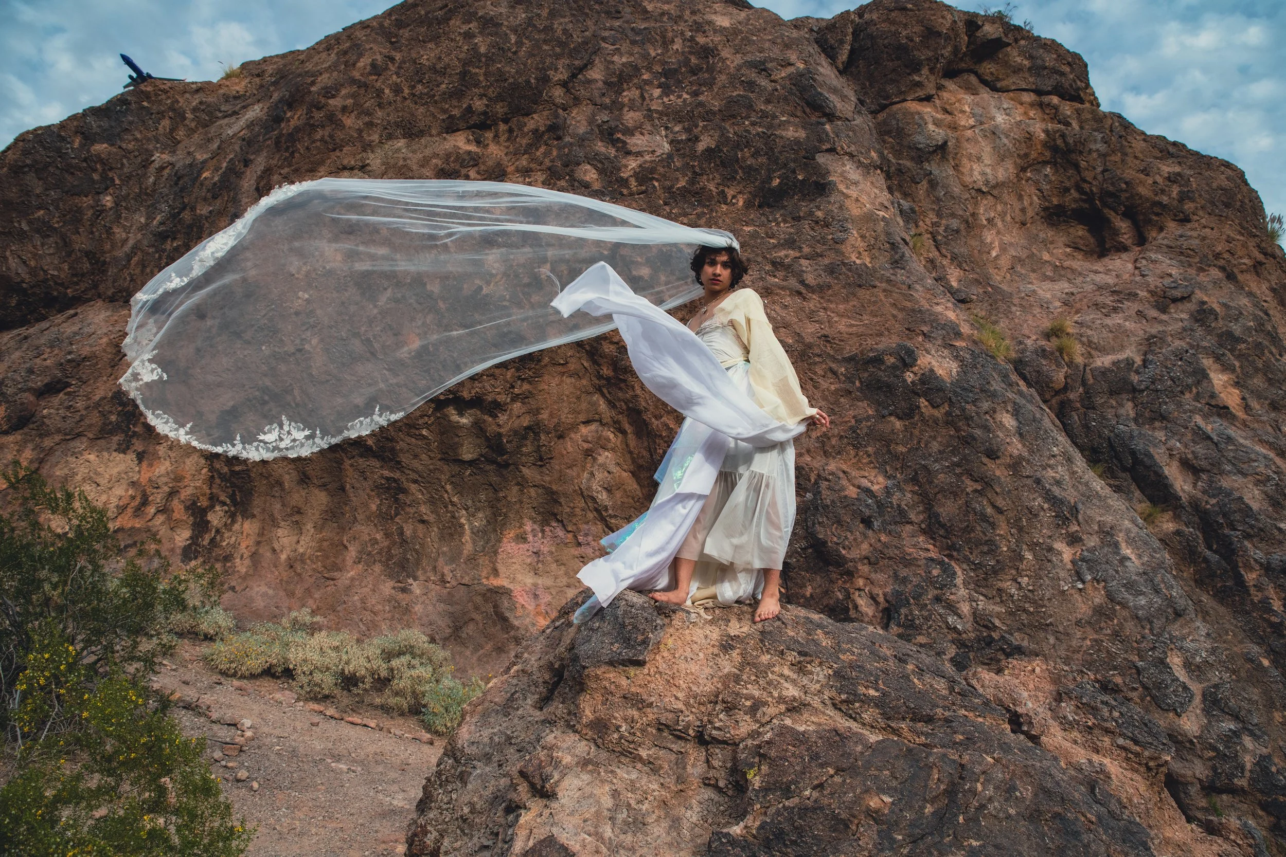 A woman with short dark hair in a white dress stands on rocks in front of a large rocky cliff, with a flowing white sheer fabric around her. An airplane is visible in the sky above the cliff.