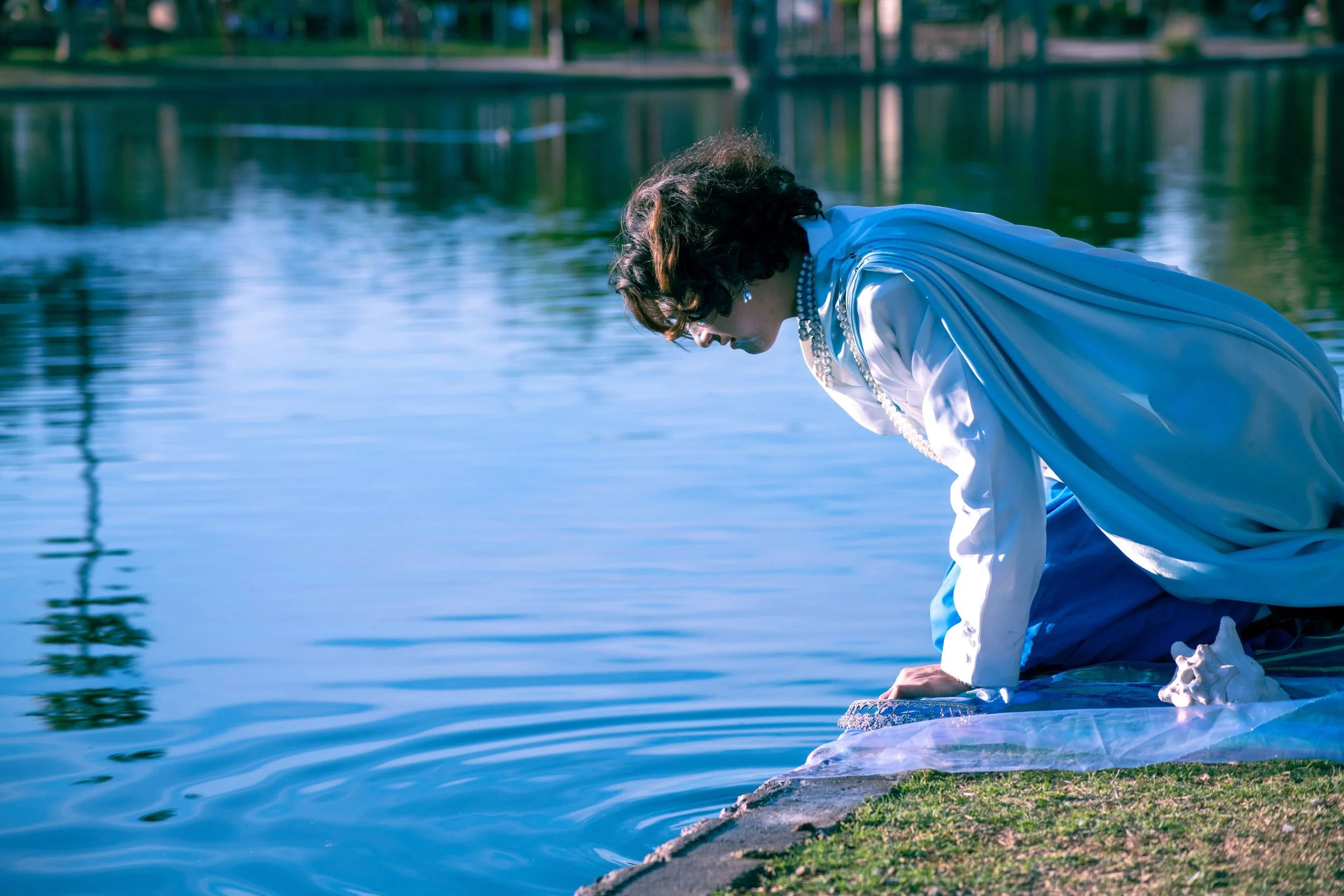 Woman in a white dress kneeling by a lake, with trees and a bridge in the background.