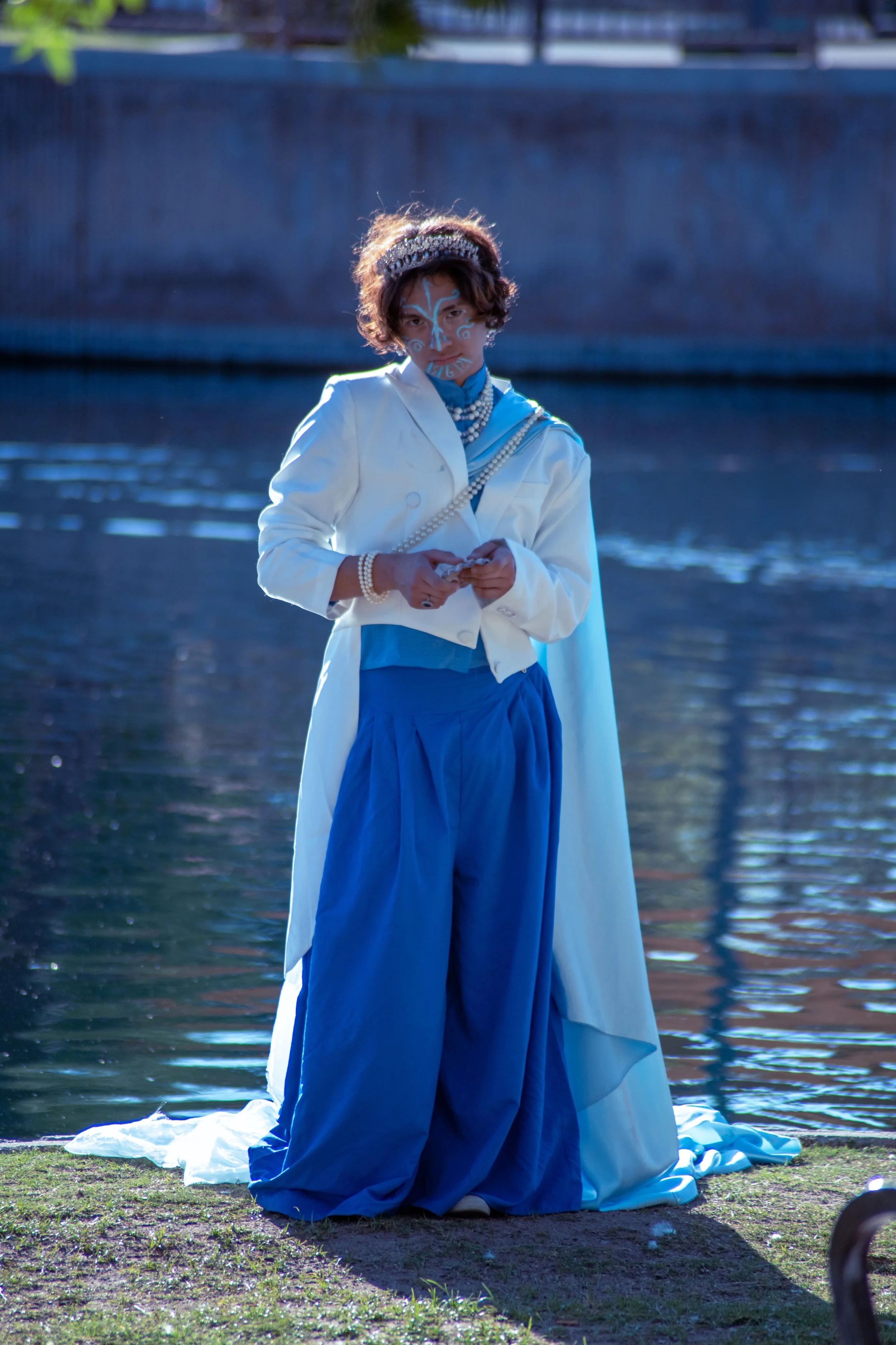A person dressed in blue and white, with face paint and jewelry, standing near a body of water in an outdoor setting.