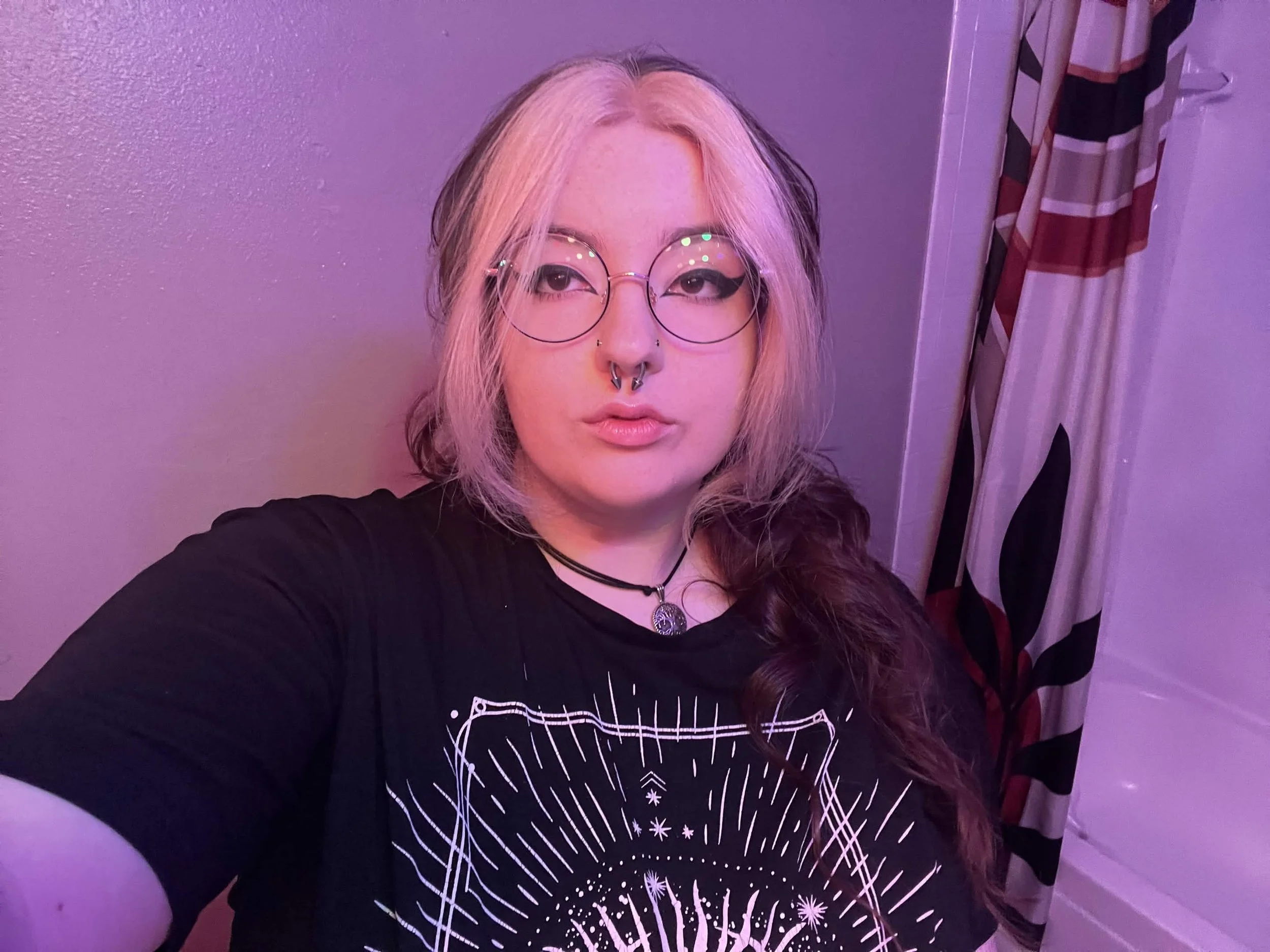 A person with long hair, wearing glasses and a septum piercing, taking a selfie against a purple-lit wall. They are wearing a black shirt with a white graphic design.