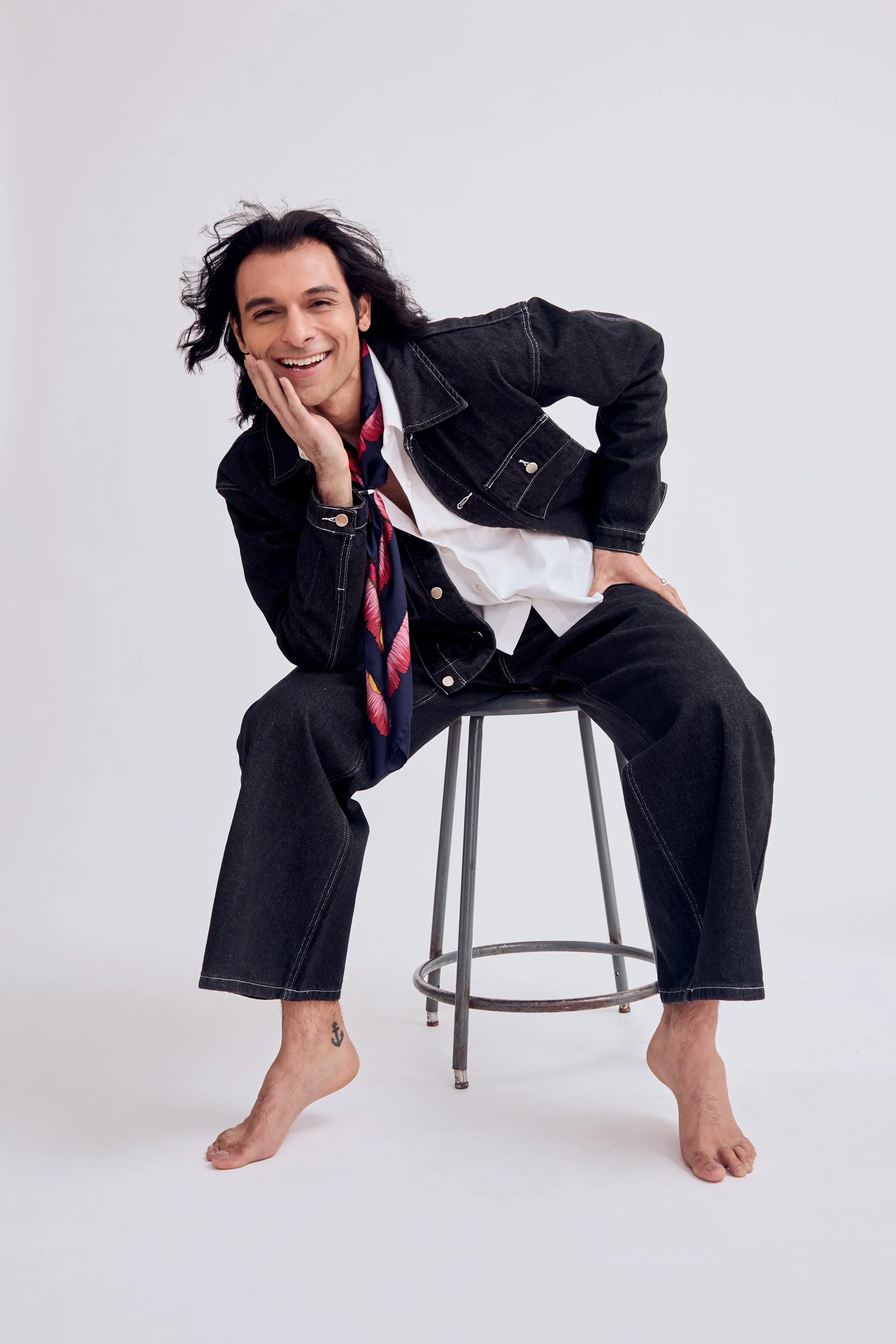 A man with long dark hair and a smile posing sitting on a stool, resting his face on his hand. He is shirtless under a black denim jacket and white shirt, with a colorful scarf around his neck, and wide-leg black jeans. He is barefoot with an anchor tattoo on his right ankle, set against a plain white background.