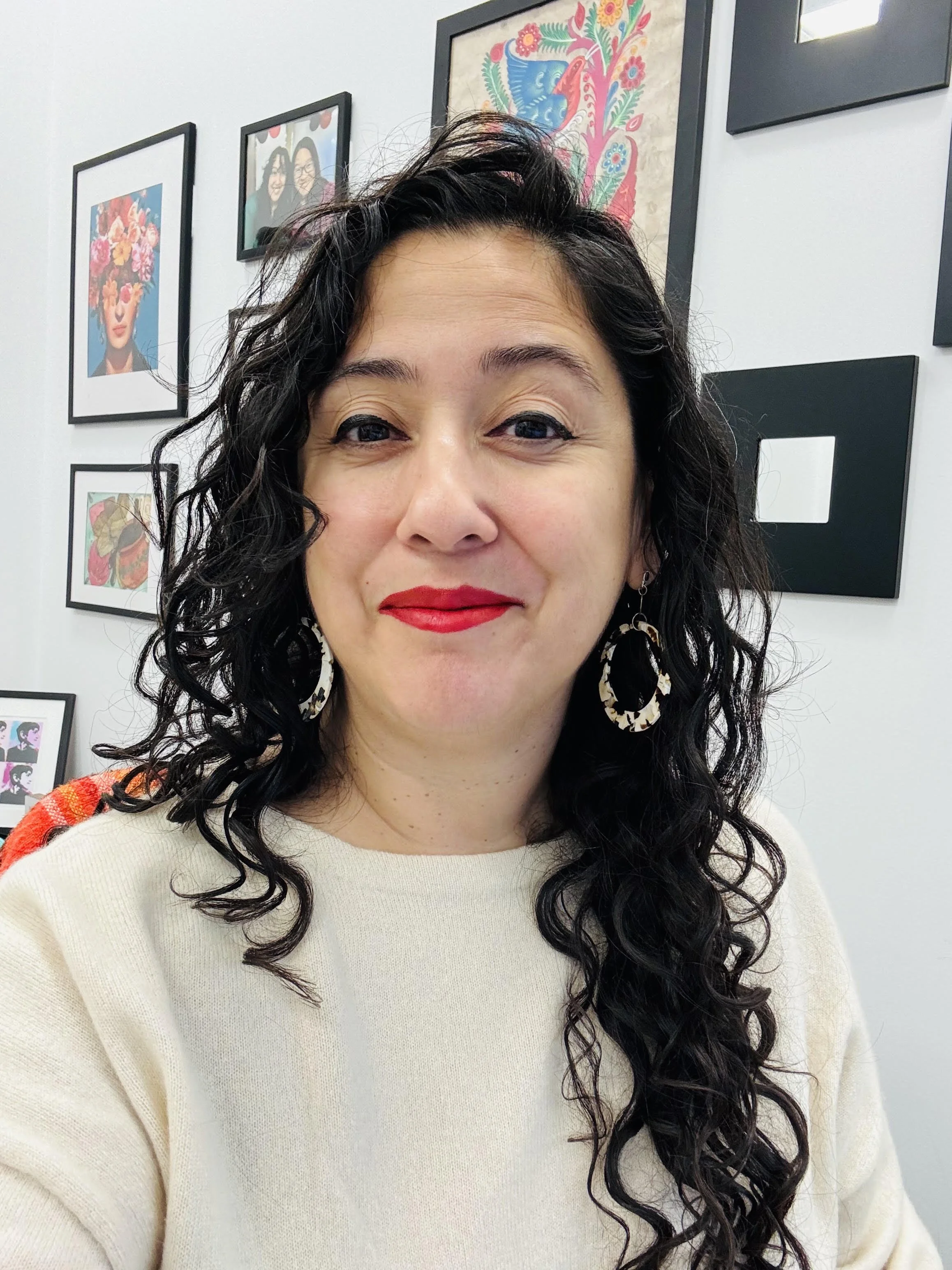 A woman with curly black hair and red lipstick, wearing a cream-colored sweater and large earrings, taking a selfie in front of a wall decorated with framed colorful artwork.