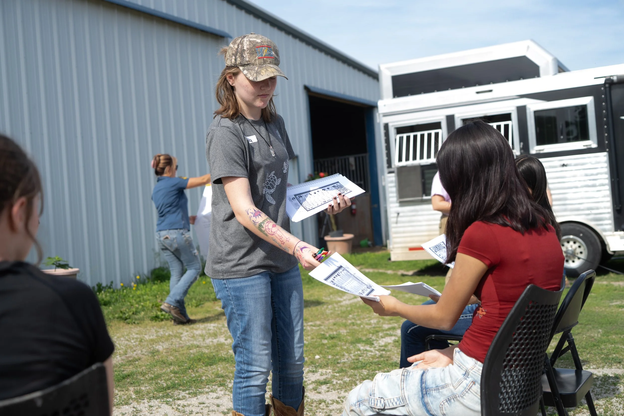 Young woman with tattoos wearing a camouflage cap and gray t-shirt handing out papers to seated people outdoors near a metal building and a horse trailer.