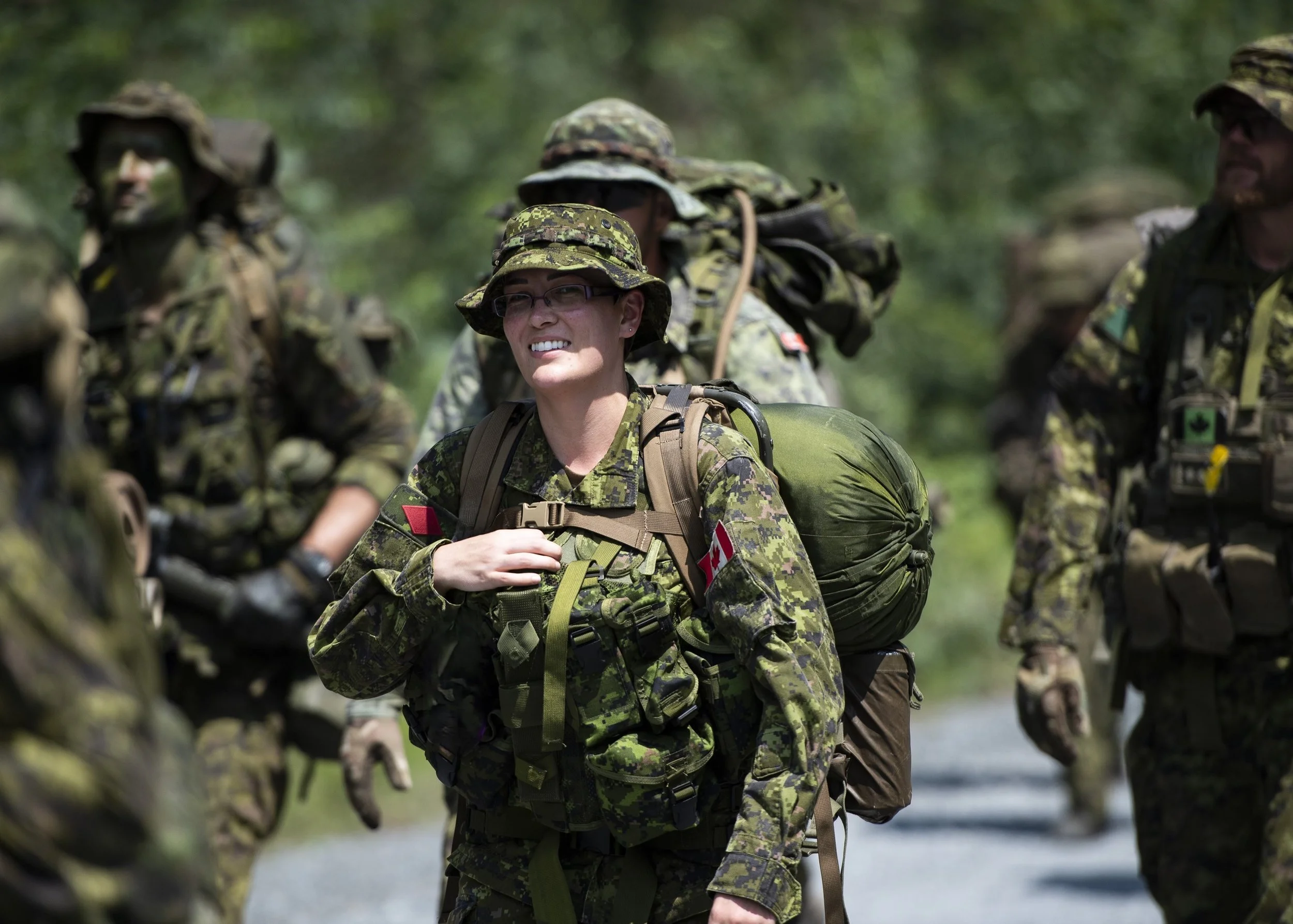 A group of soldiers in camouflage uniforms walking outdoors with backpacks and gear, one woman smiling and wearing glasses.