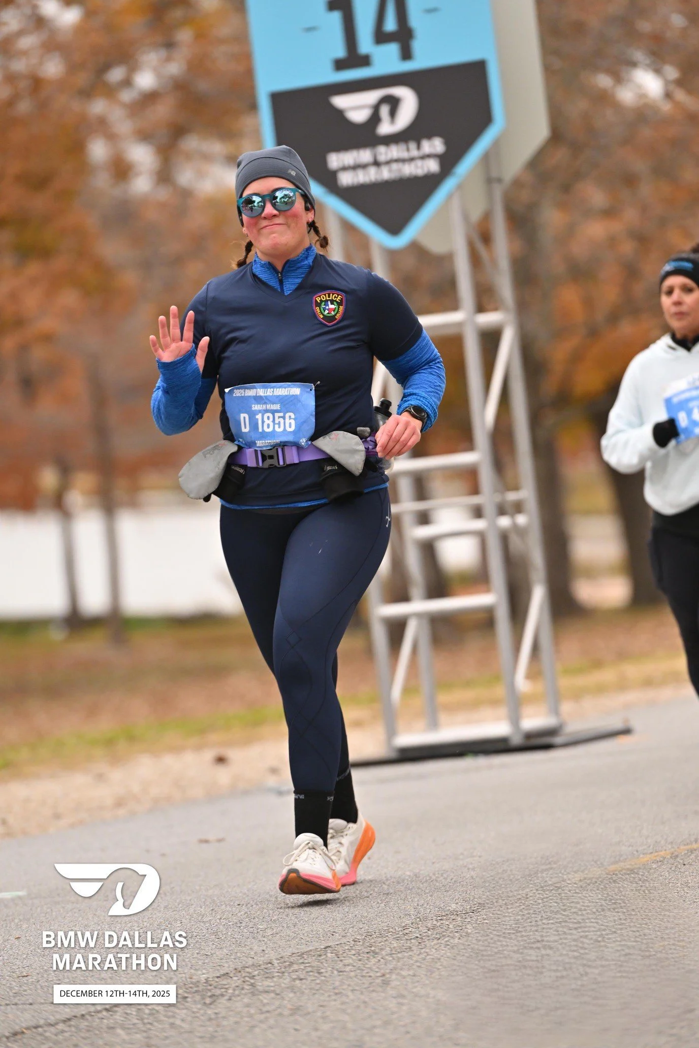 A woman running in a marathon, wearing sunglasses, a beanie, a water bottle holder, and a blue race bib with the number 1856, participating in the BMW Dallas Marathon held from December 14th, 2025 in Dallas, Texas.