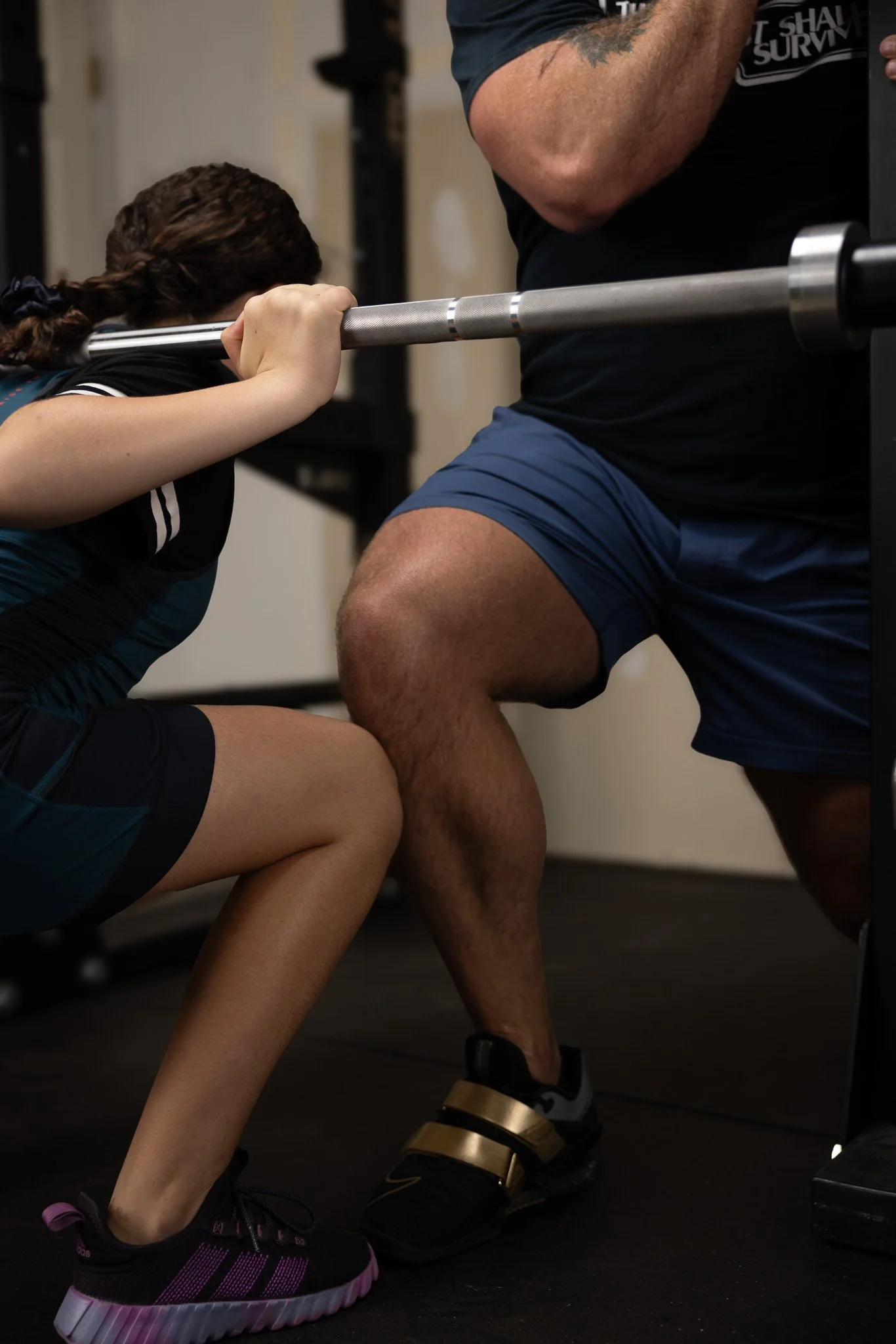A young woman is squat exercising with her hand on a barbell, supervised by a man, in a gym setting.