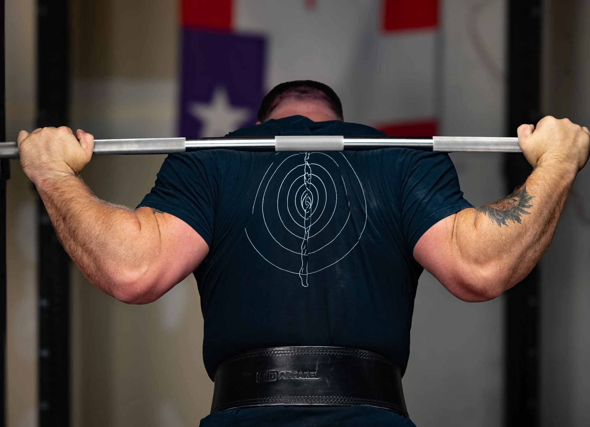 A man with a tattoo on his left arm is preparing to lift a barbell in a gym, with his back facing the camera. The focus is on his muscular back and arms, and he is wearing a black t-shirt.