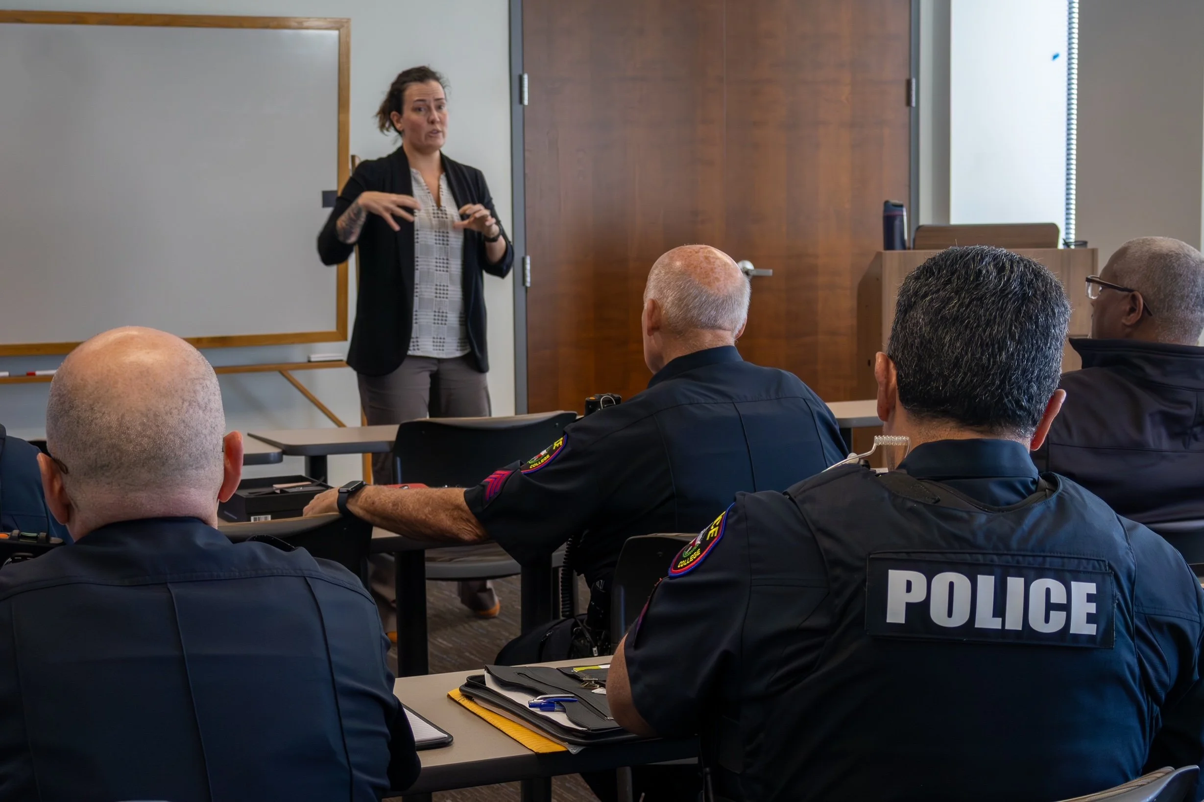 A woman is standing and speaking to a group of police officers sitting at tables in a training room. The officers are wearing dark uniforms with police labels on their backs. The room has a whiteboard, a wooden door, and a window with blinds.