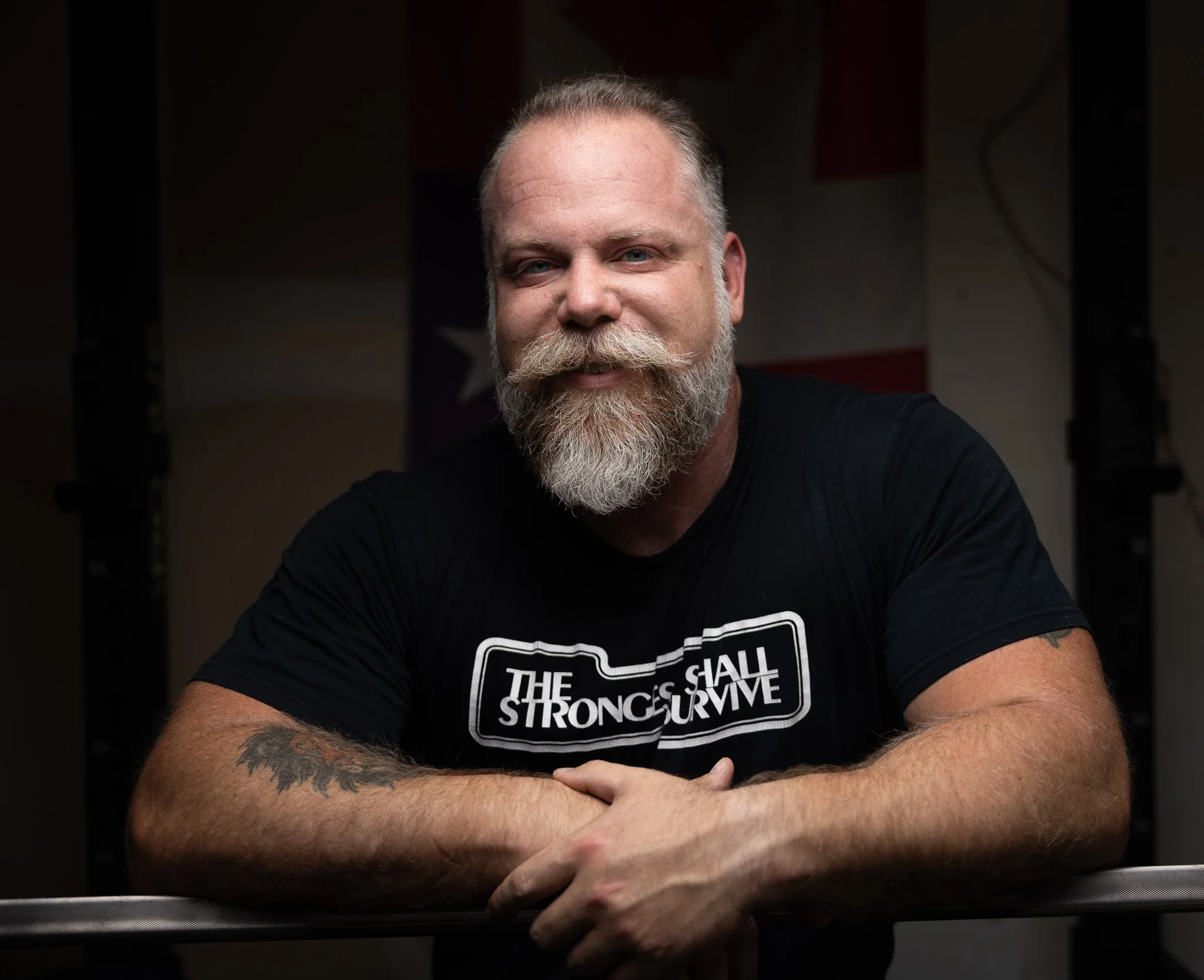 A man with a beard and mustache smiling, wearing a black T-shirt with text, sitting with arms crossed, in front of a dark background.