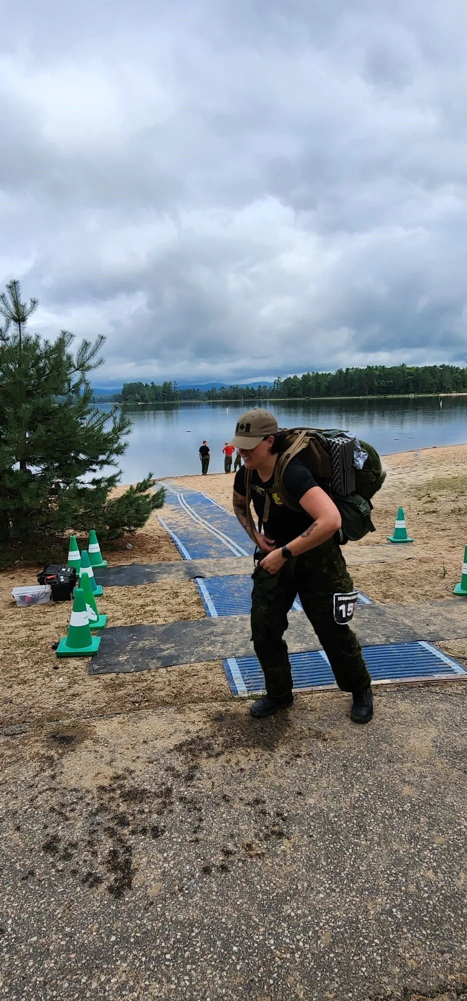 A woman in military uniform and a cap, carrying a large rucksack, standing on a boat ramp near a lake, with a person in black in the background and a cloudy sky overhead.