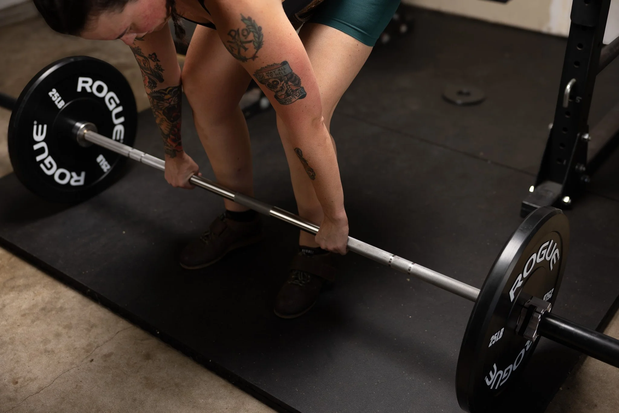 A person with tattoos is bending over to lift a loaded barbell on a black rubber mat in a gym. The person is wearing glasses and dark workout clothes. The gym equipment includes weight plates and a squat rack.