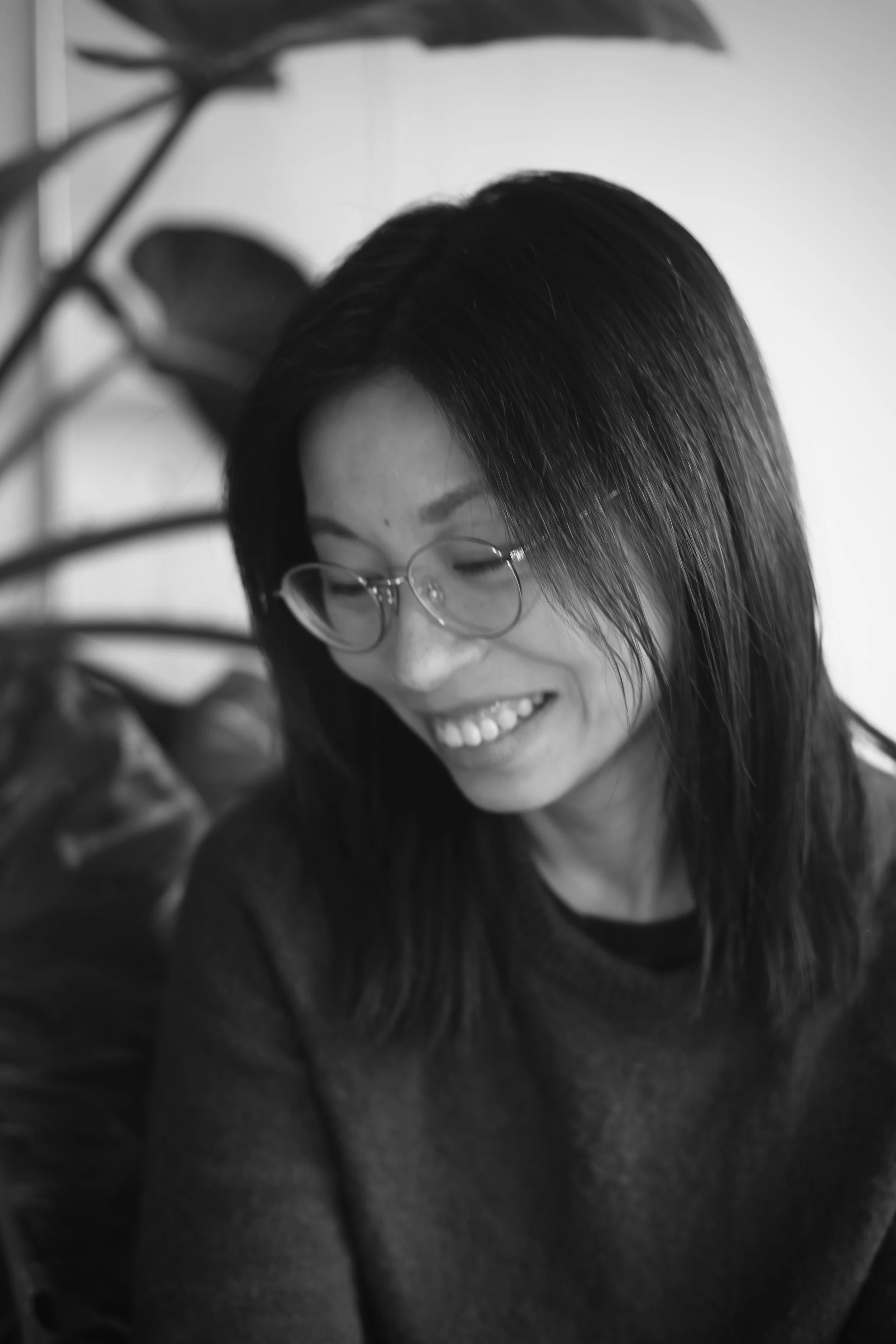 Black and white photo of a person with long dark hair, glasses, and a bright smile, sitting indoors near a plant.