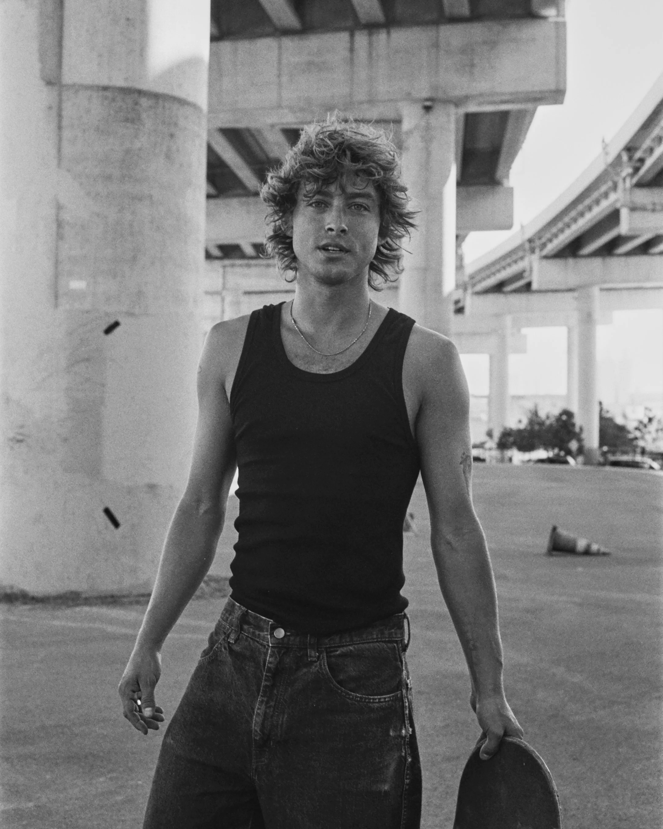 A black-and-white photo of a young man with curly hair, wearing a black tank top and jeans, standing outdoors under a bridge or overpass, holding a skateboard in his left hand.