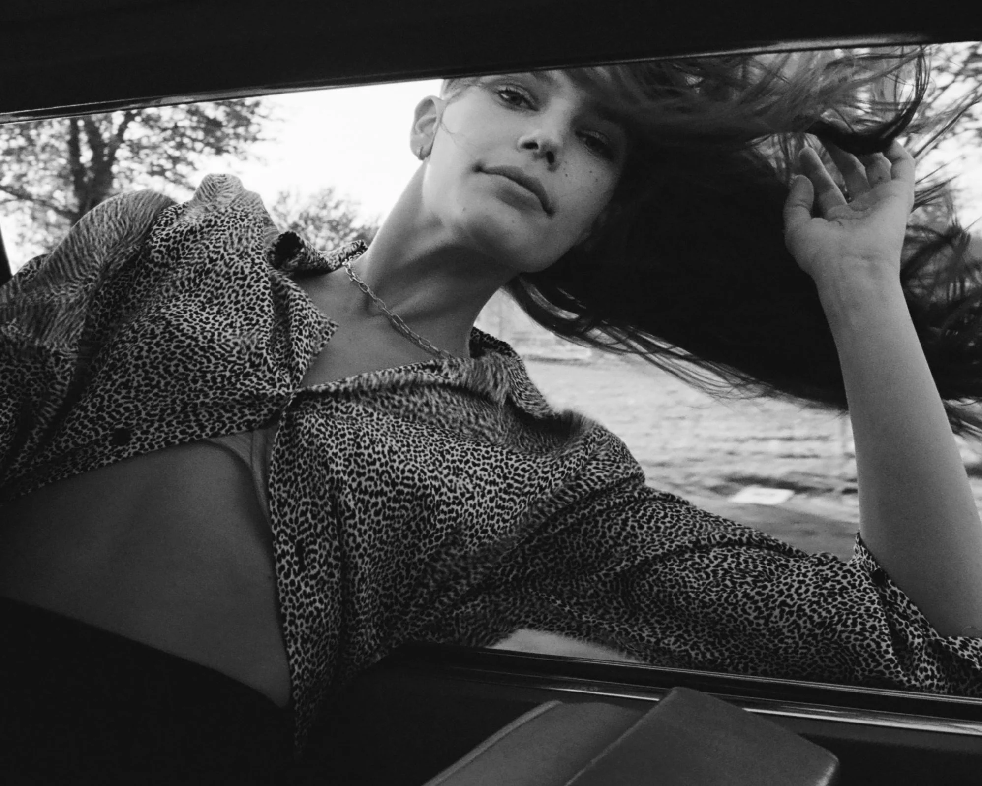 Black and white photo of a woman with long hair lying down, wearing a patterned top and a chain necklace, with trees visible in the background.
