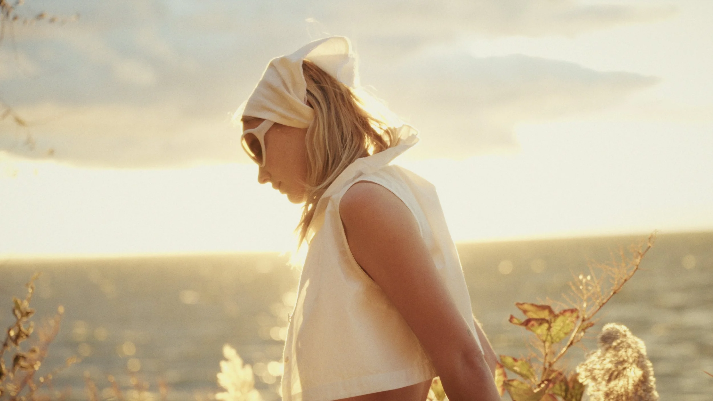 A woman with blonde hair wearing a sleeveless dress and a headscarf, standing outdoors during sunset, with plants and a blurred natural background.