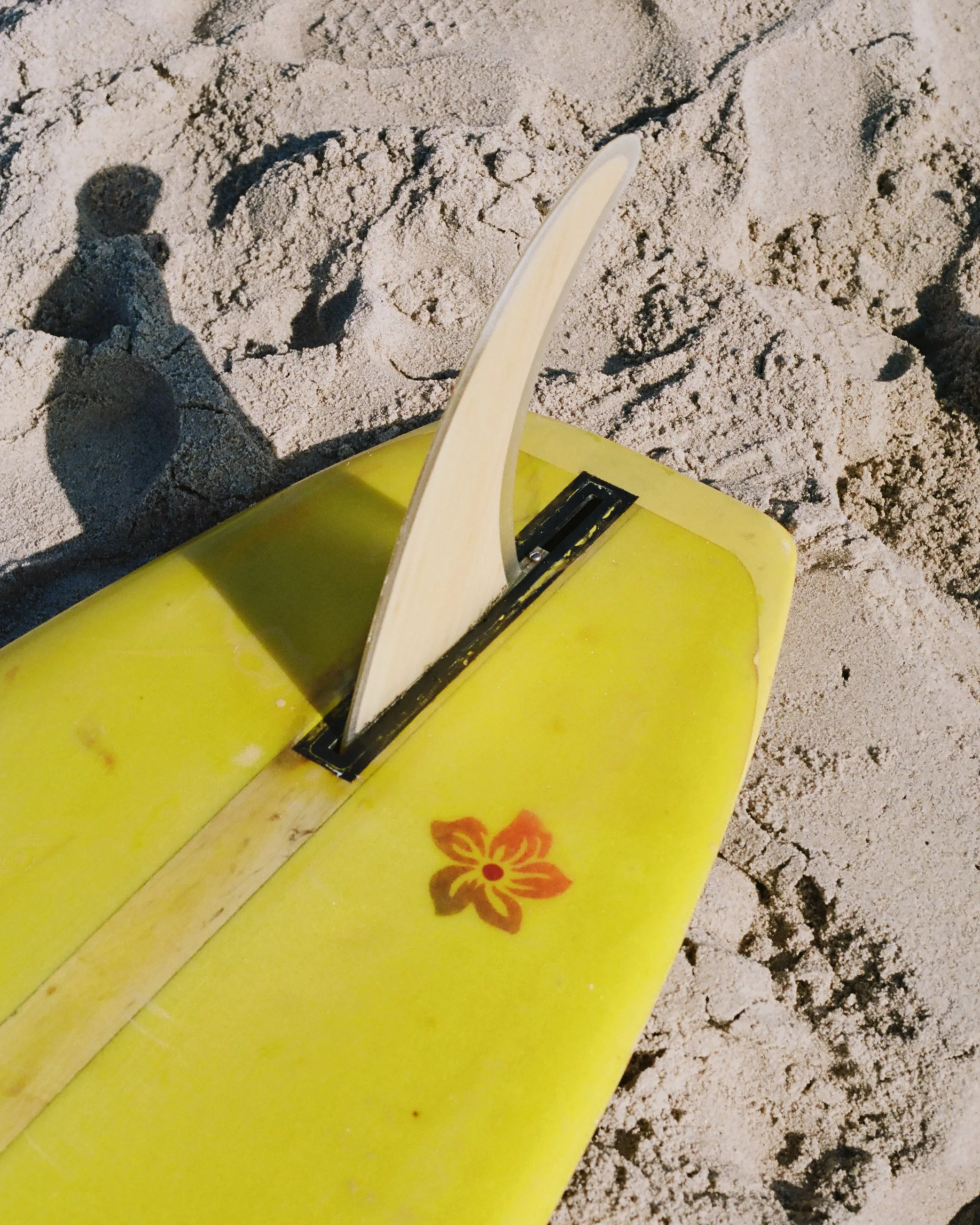 A yellow surfboard with a pink flower design lying on sandy ground, with a wooden and metal object resting on it, casting shadows on the sand.