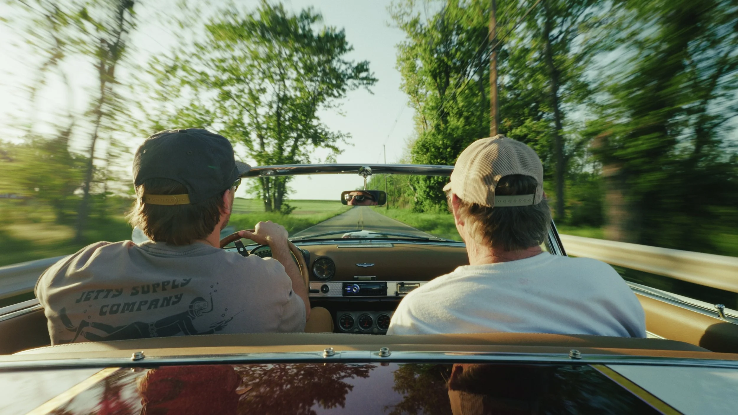 Two young men in hats driving a vintage convertible along a rural road surrounded by trees and green landscape in sunlight.