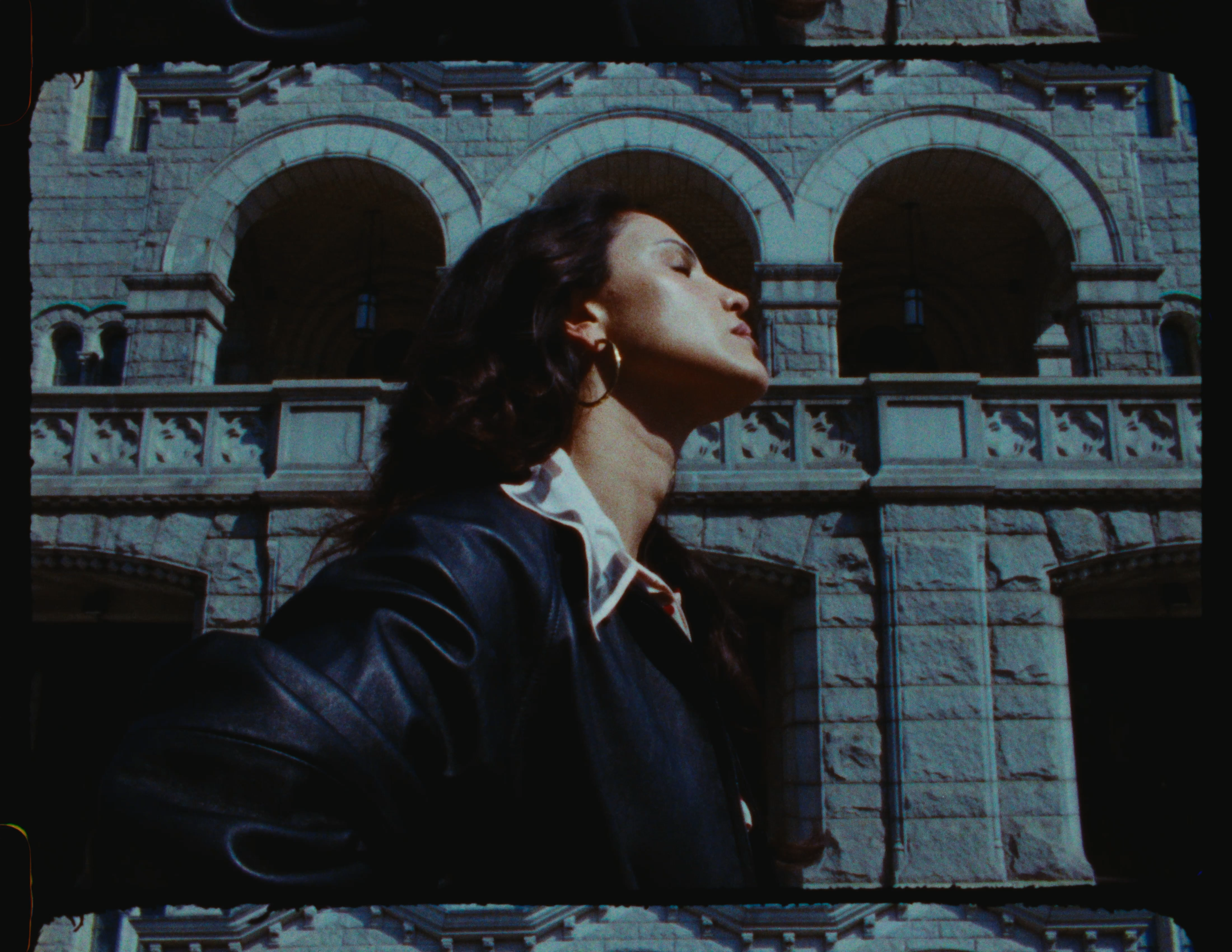 A woman with dark curly hair wearing earrings and a leather jacket stands in front of a historical stone building with arches.