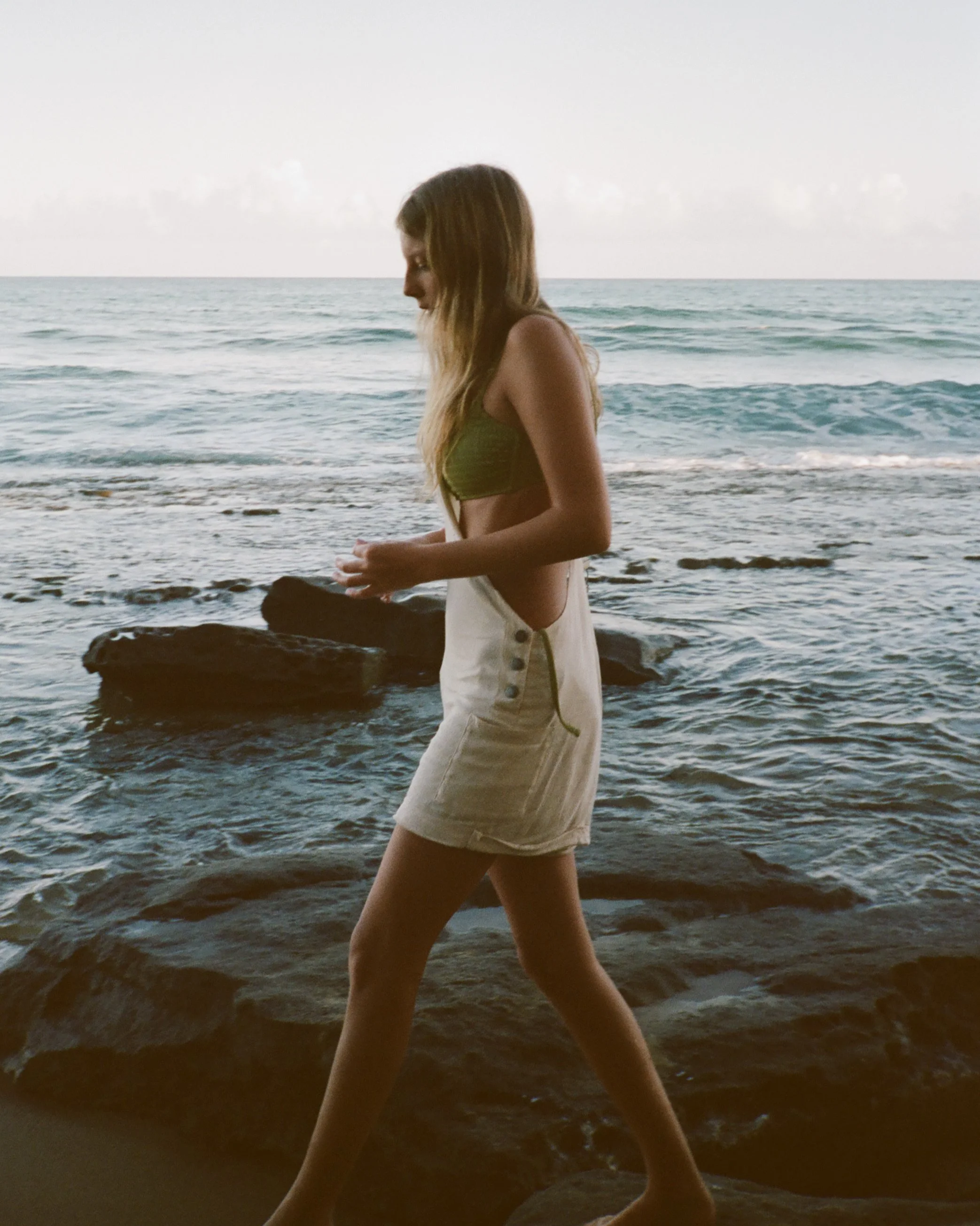 A woman walking along a rocky beach at sunset or sunrise with the ocean in the background.