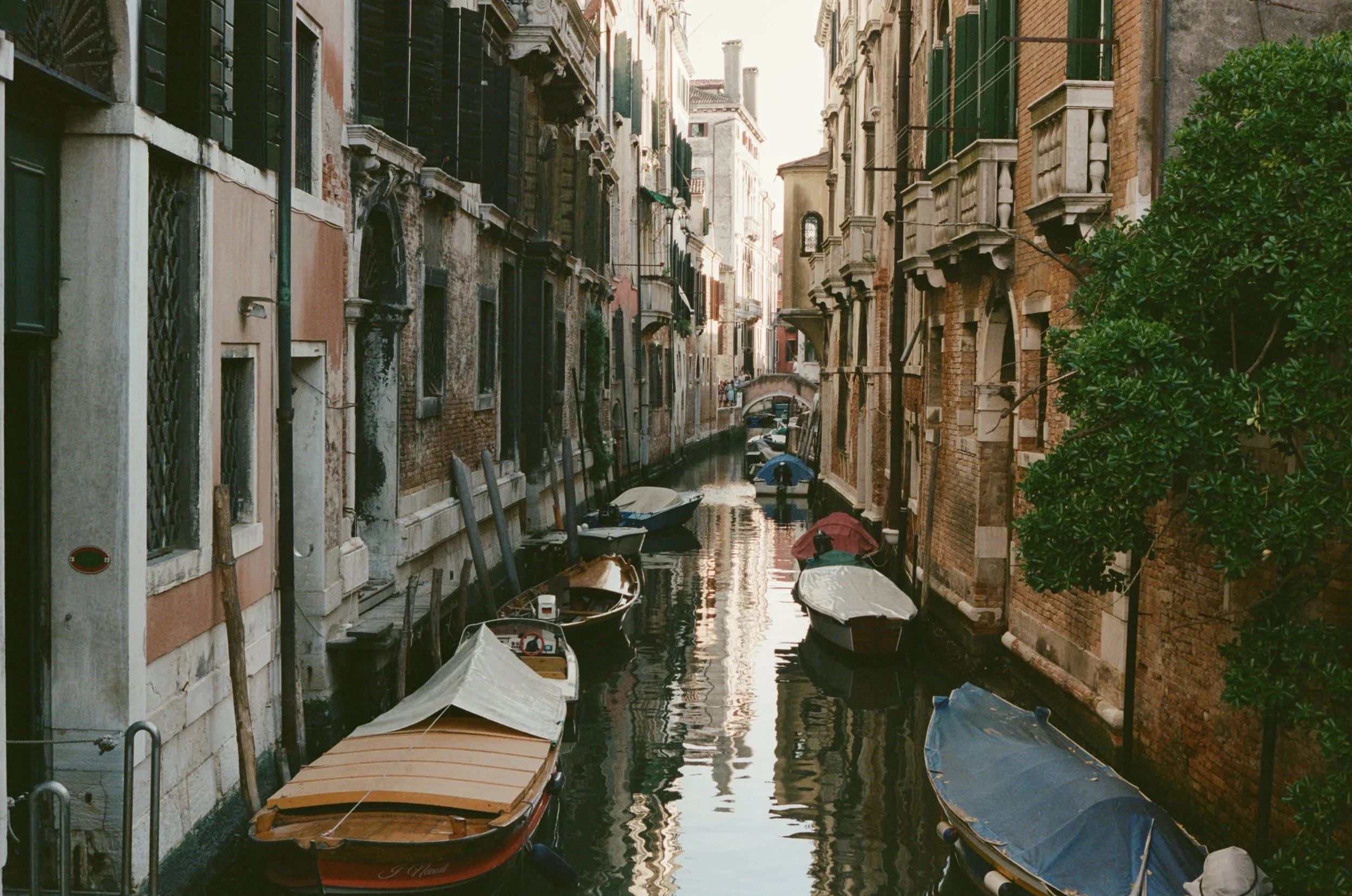 A narrow canal in Venice, Italy, with several boats docked along the sides, bordered by old, weathered buildings with small balconies and green shutters, and a person walking in the distance under an umbrella.