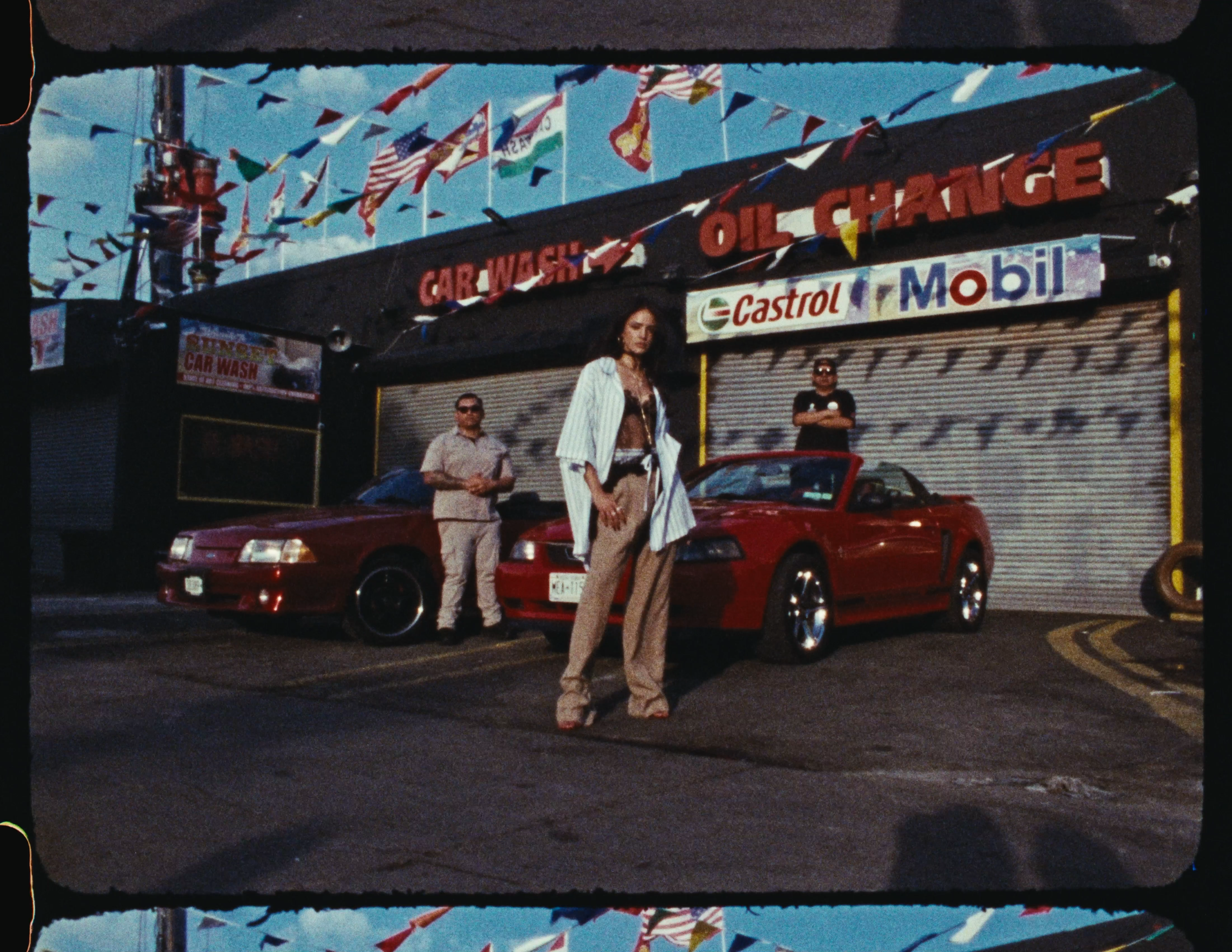 People standing in front of a car wash shop with colorful flags and automotive brand signs, including Castrol and Mobil, under a blue sky.