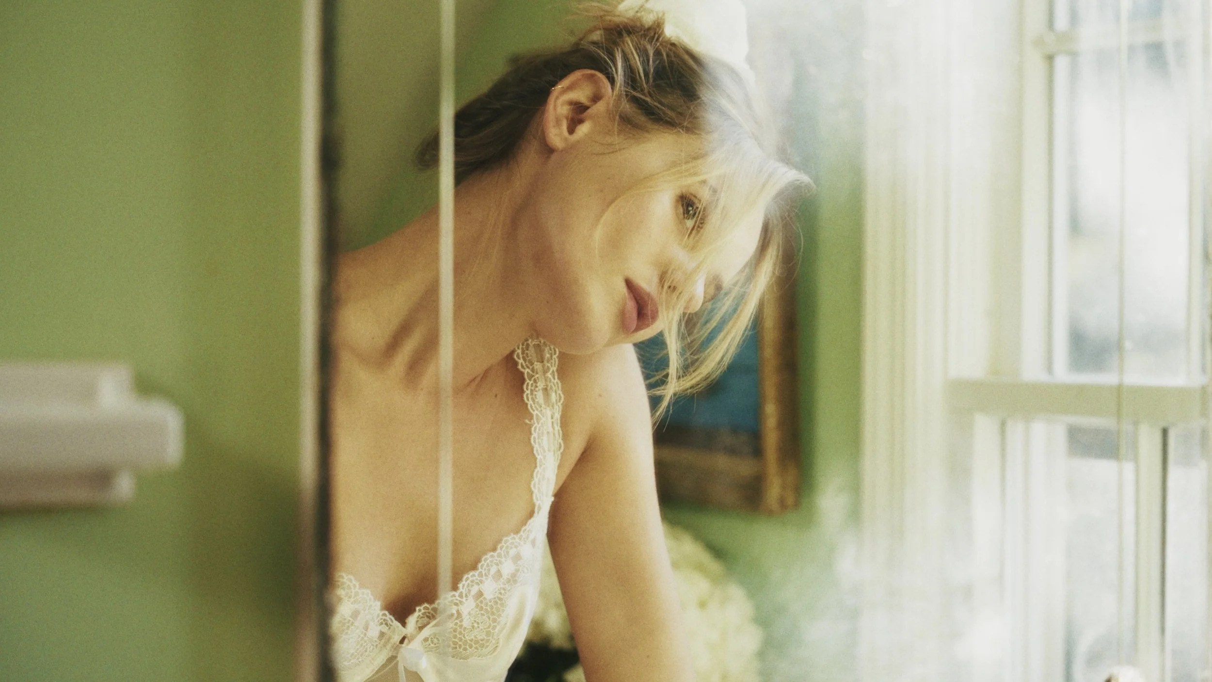 A woman with blonde hair looking out the window, wearing a white lace top, in a room with green walls and a window with white curtains.